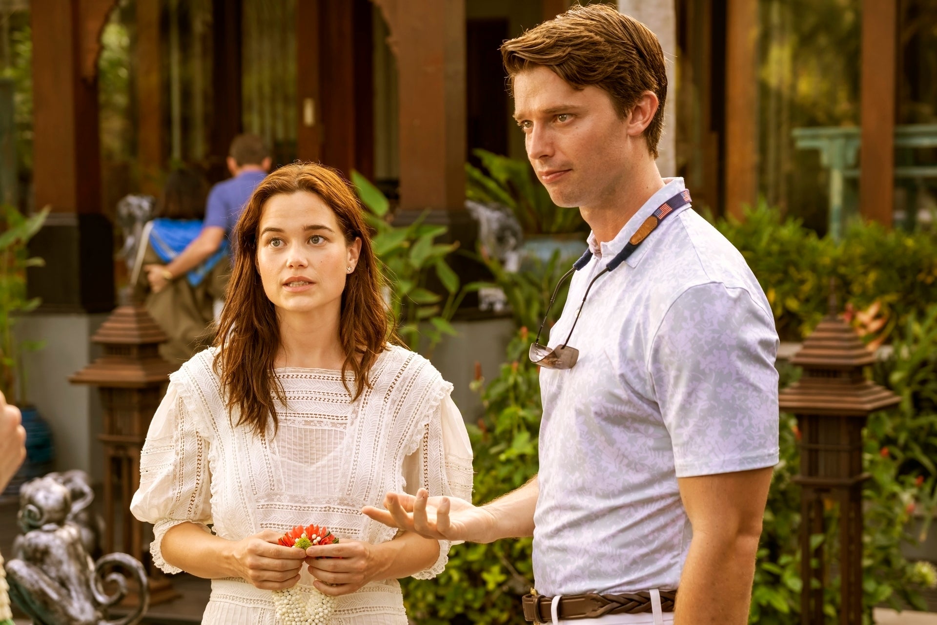 Two actors stand outside on a set. The woman wears a delicate, white lace dress. The man is in a casual polo shirt with sunglasses