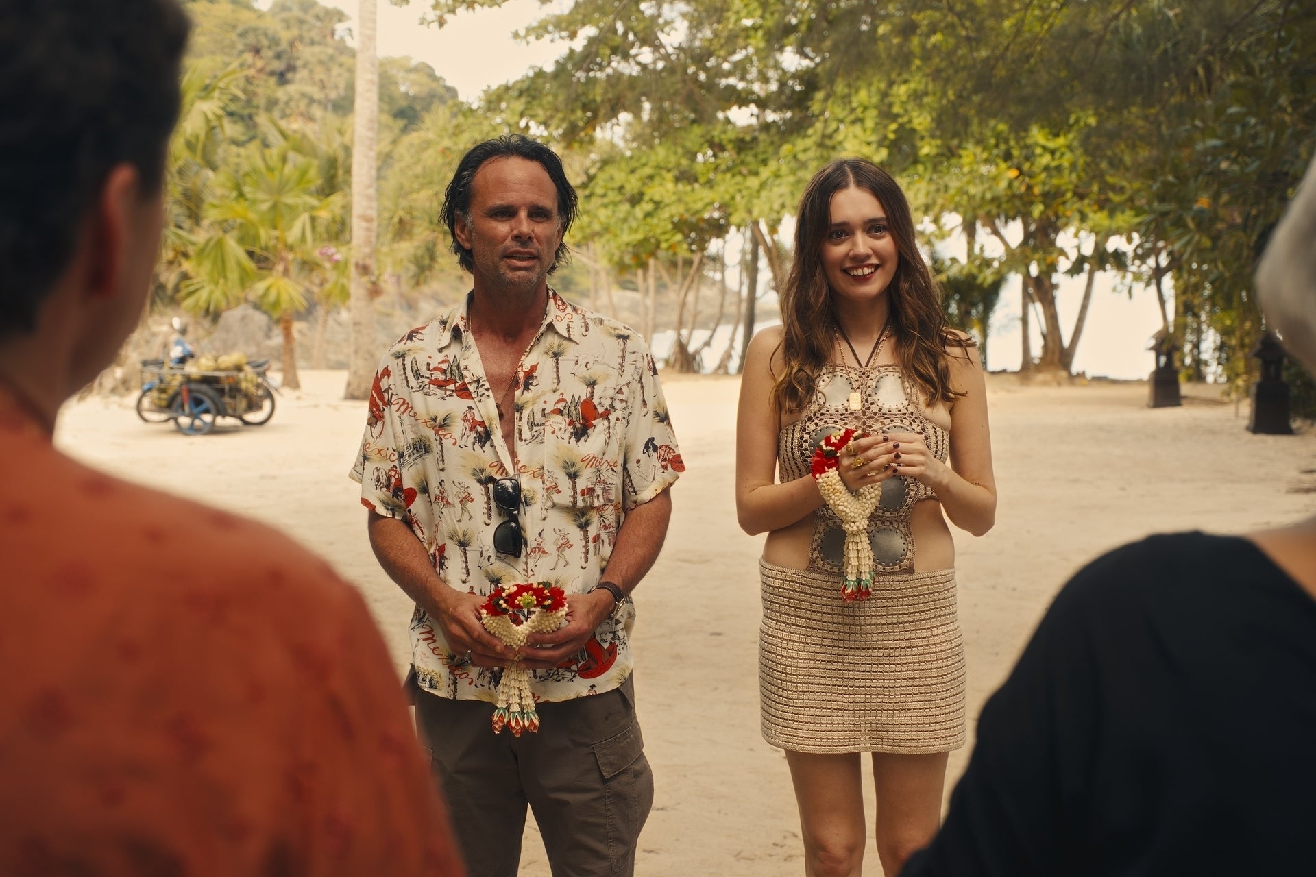 A man in a patterned shirt and a woman in a crocheted outfit stand outdoors, holding floral items, engaging with people out of view