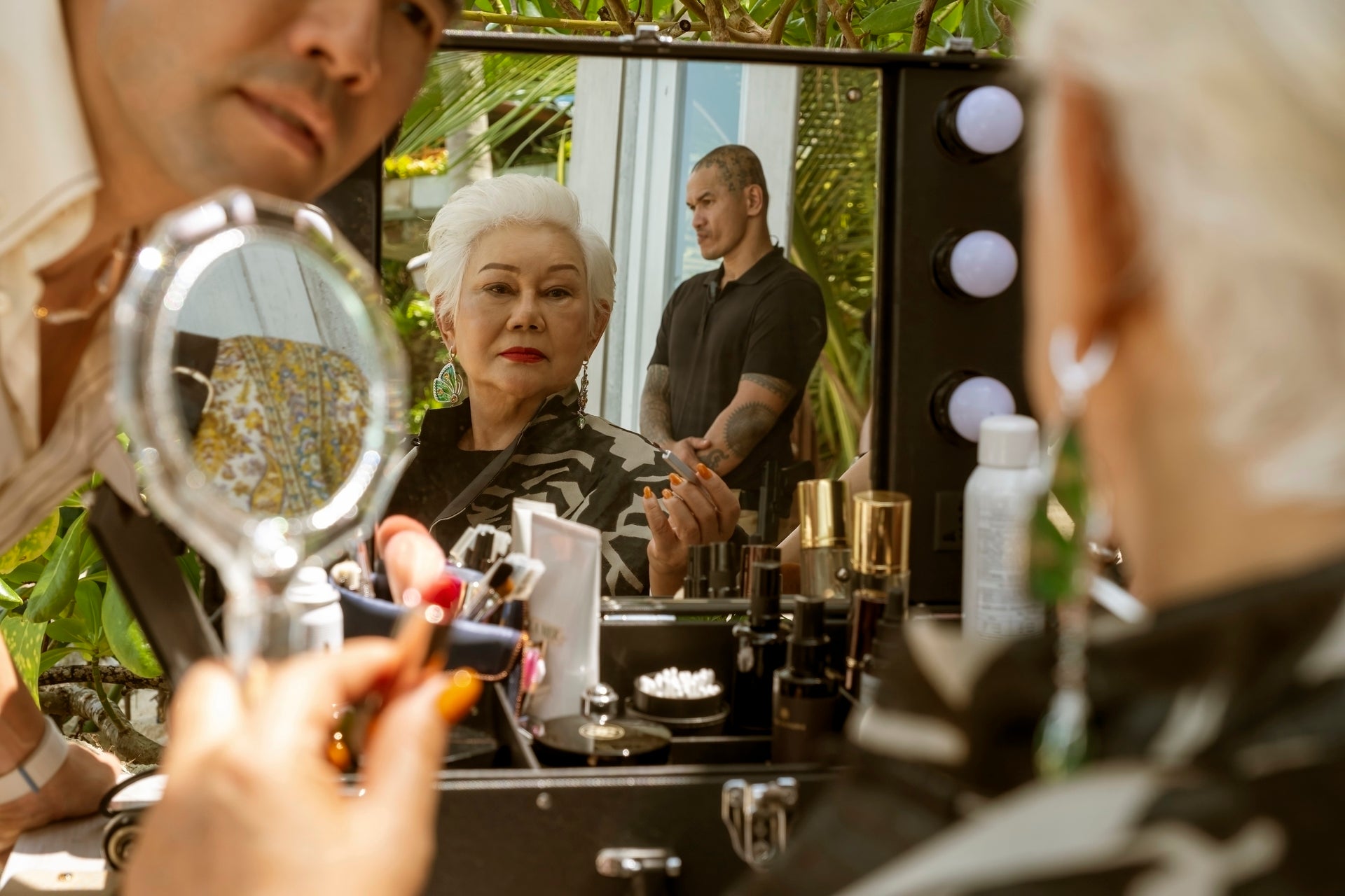Person with short hair looks in a mirror during a makeup session outdoors, with a person in the background and beauty products on the table