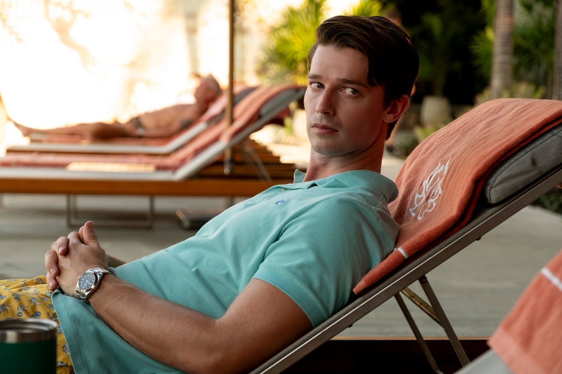 Person lounging on a deck chair by a pool, wearing a polo shirt and patterned shorts, looking at the camera