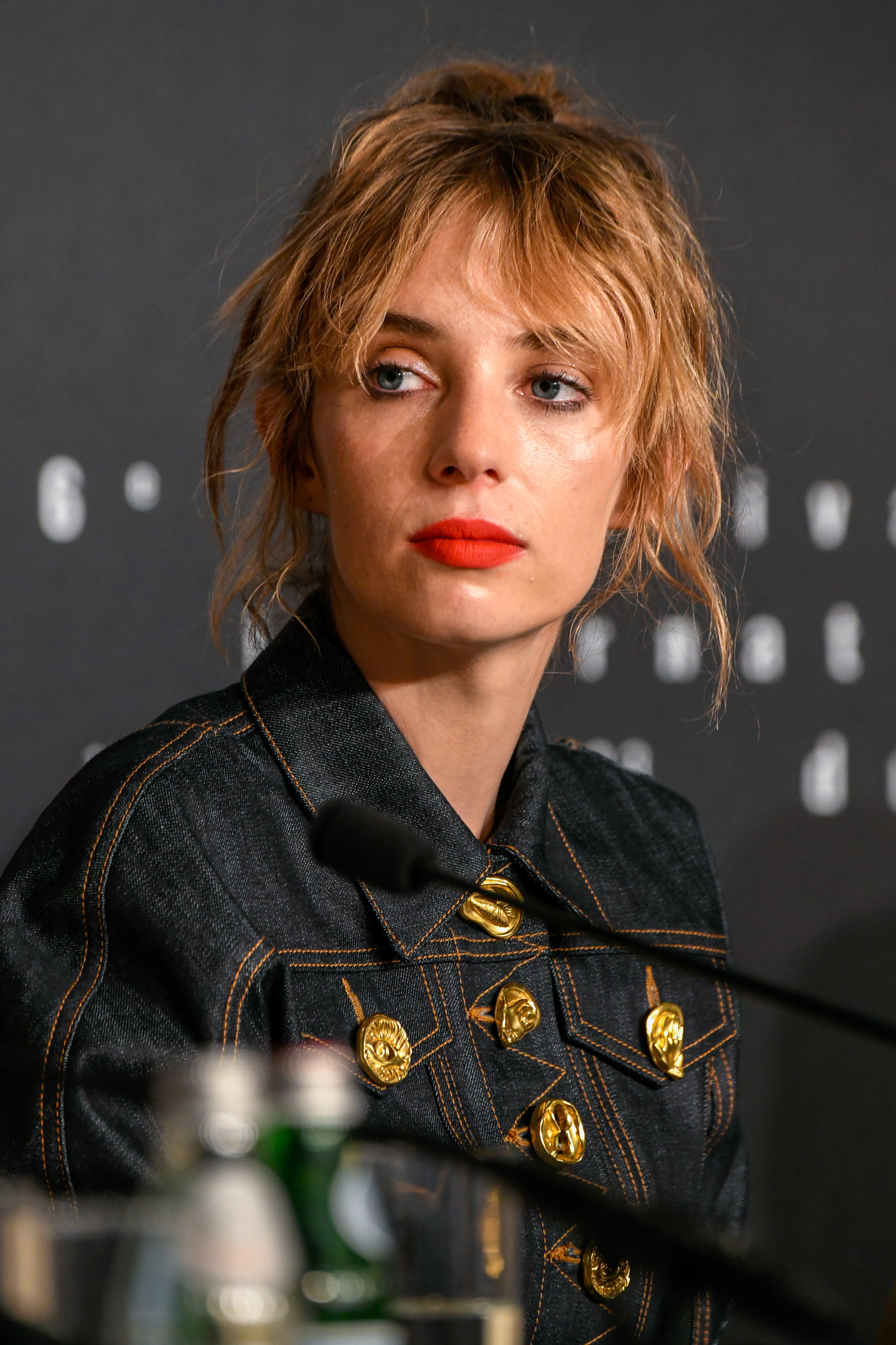 Maya Hawke with tousled hair in a denim jacket with buttons, seated at a press event