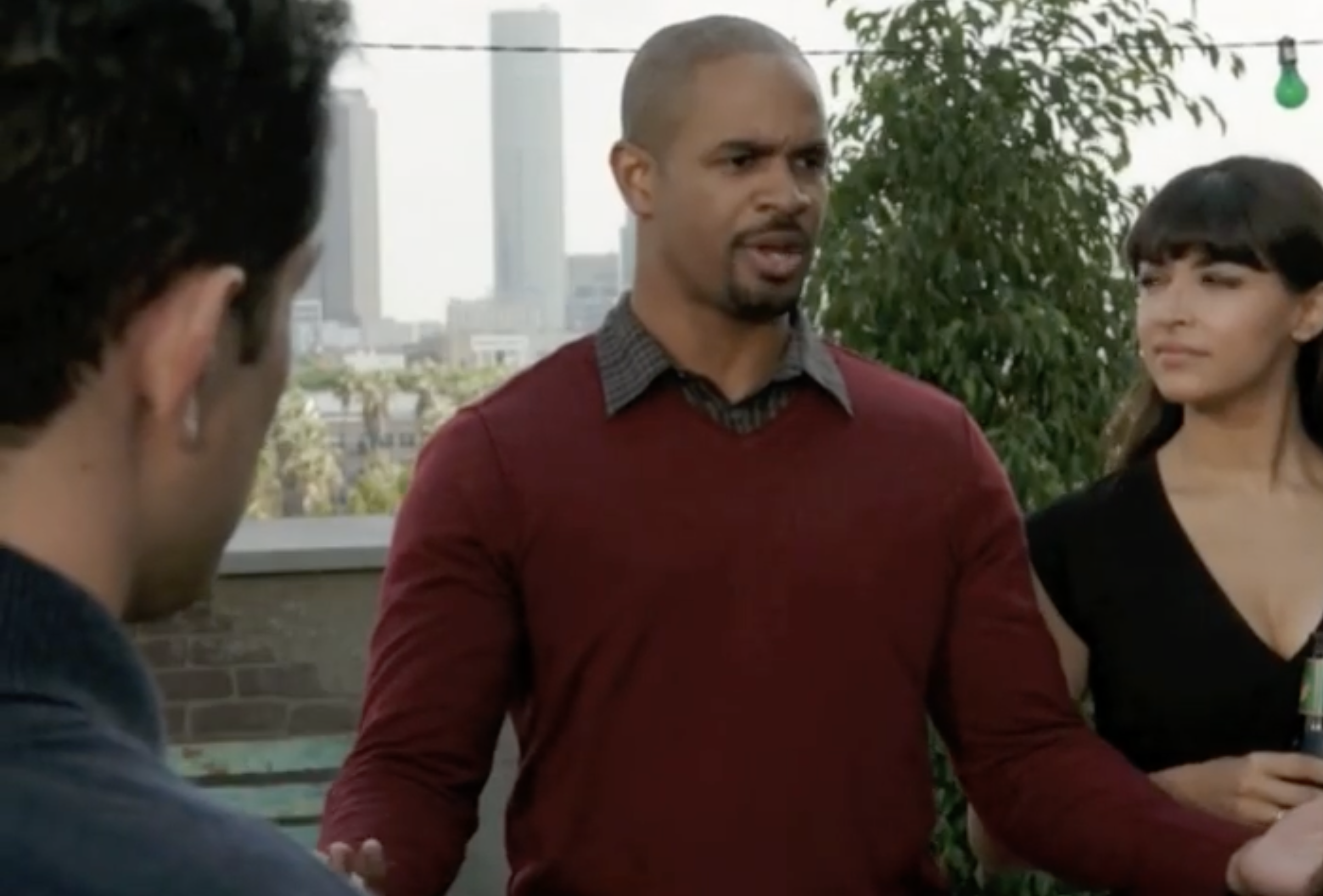 A man in a red sweater and a woman in a black dress converse with a third person on a rooftop, with a city skyline in the background