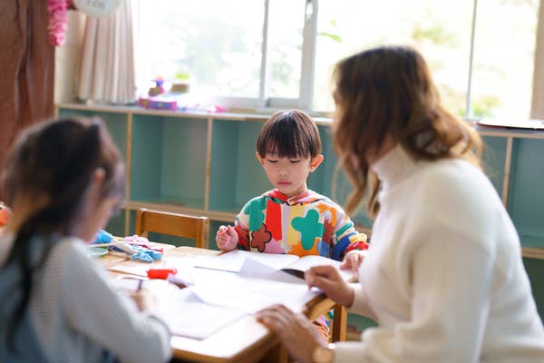 A teacher guides two young children working on crafts at a table in a classroom