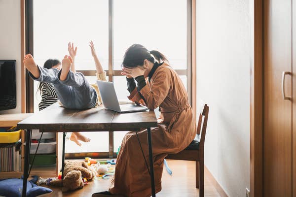 A person in a robe looks stressed at a table with a laptop; two children play energetically nearby, toys scattered on the floor