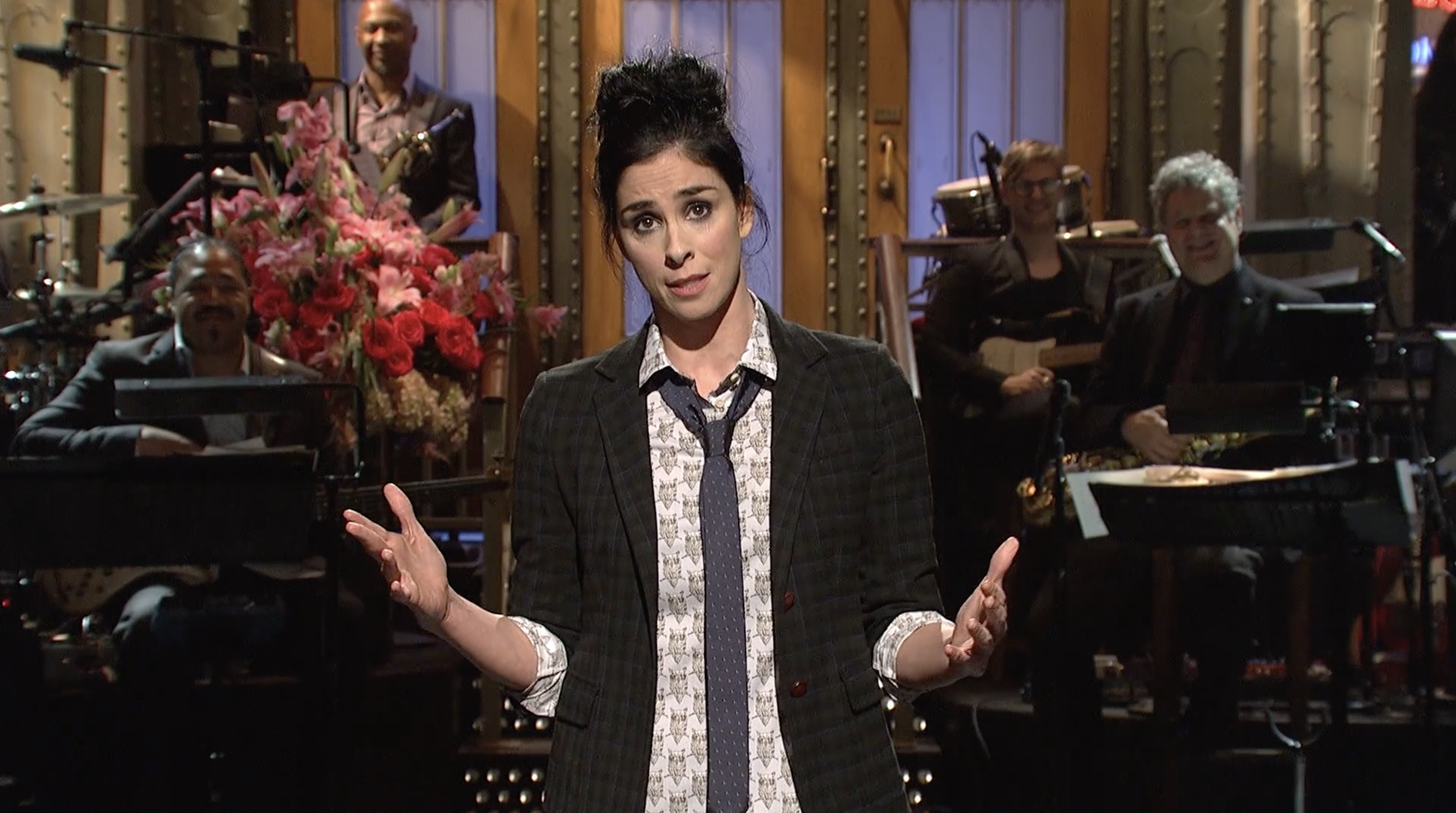 A person stands on a stage with a band in the background, wearing a checkered suit jacket and a loosely tied necktie, gesturing expressively