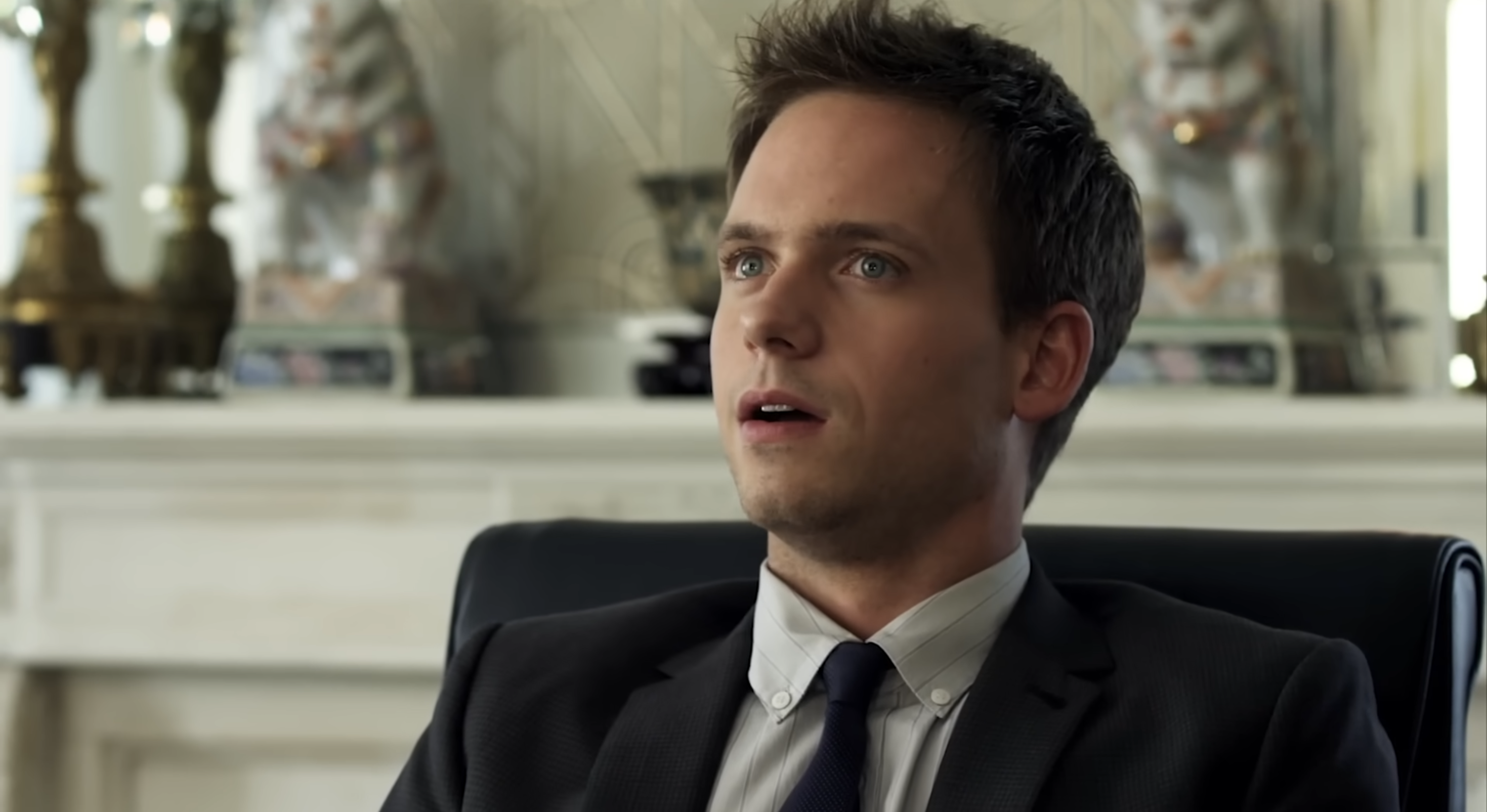 Person in a suit sitting in a chair, appearing surprised, with decorative items in the background
