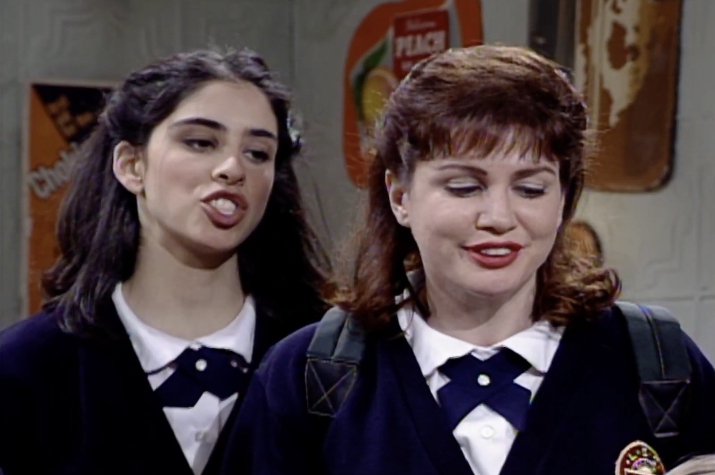Two women in school uniforms are standing indoors, one smiling with a backpack. Posters are visible in the background