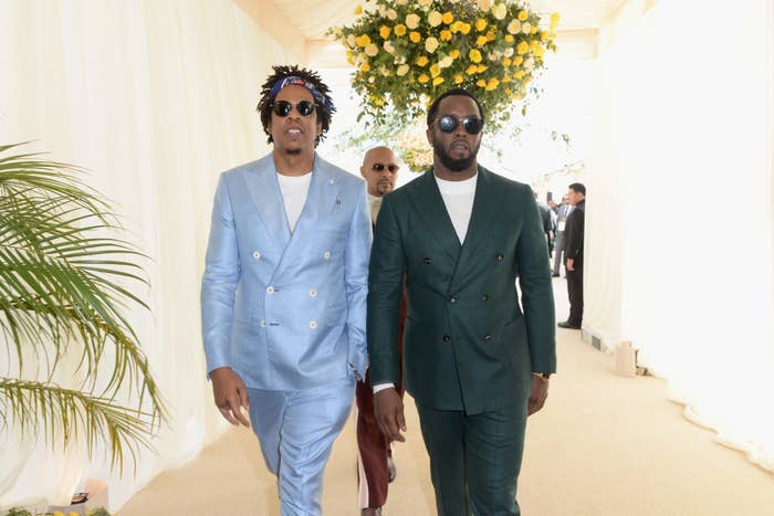 Jay-Z and Diddy in stylish suits walk together at an event. One wears a double-breasted light suit, the other a dark suit. Both have sunglasses