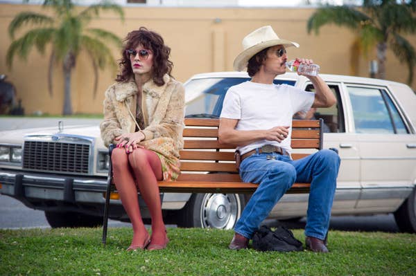 Two people sit on a bench; one wears a fur coat and sunglasses, the other a cowboy hat and jeans, drinking water. A vintage car is parked behind them