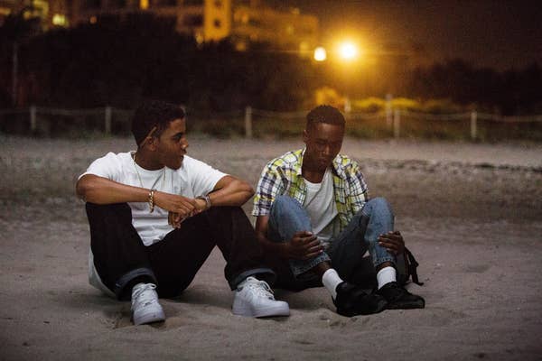 Two individuals sit on a beach at night, casually dressed, engaged in a conversation, with city lights in the background