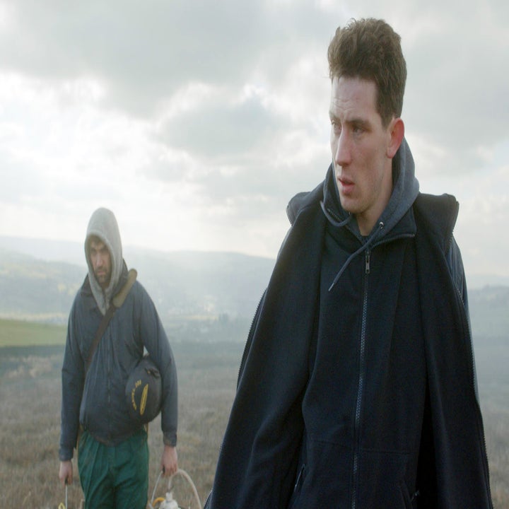 Two individuals walk outdoors in a rural setting wearing casual layers. One carries sports equipment. The mood is contemplative
