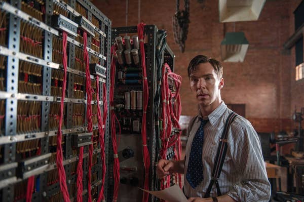 Person in suspenders and tie stands by a vintage computer with red cables, depicting a historical or film scene about codebreaking