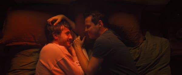 Two people are lying in bed, facing each other and smiling, sharing an intimate moment