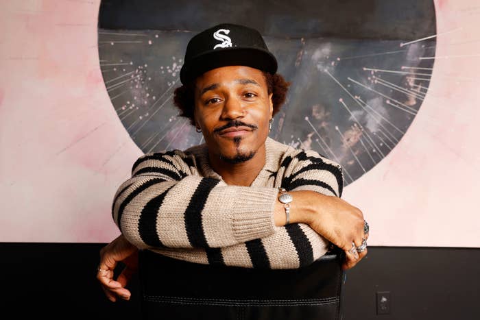 Person in a striped sweater and baseball cap leans on a chair, smiling slightly, with a geometrical mural background
