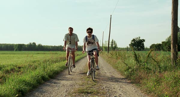 Two people leisurely biking along a rural path surrounded by greenery and open fields on a sunny day