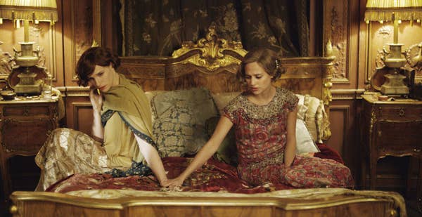 Two individuals in elegant, period-style clothing sit on a lavishly decorated bed, conveying a sense of introspection and connection