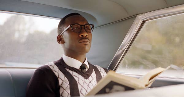 Person in glasses and a sweater vest sitting in a car, reading a book