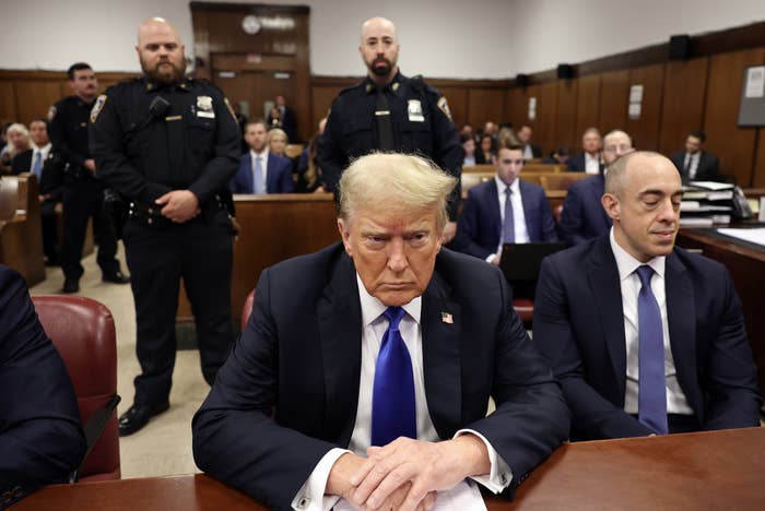 A man in a suit sits in a courtroom filled with people, with security personnel standing behind