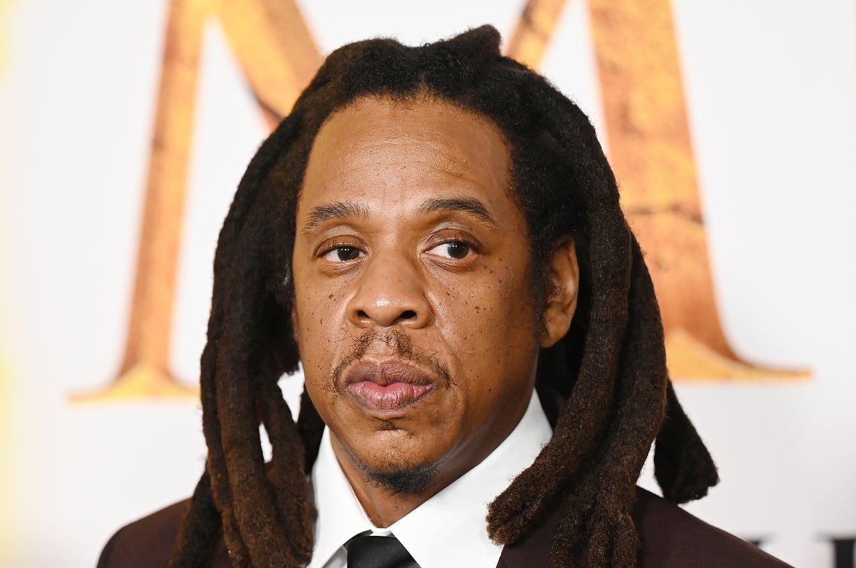 Jay-Z with shoulder-length dreadlocks wears a suit and tie, attending an event with a patterned backdrop