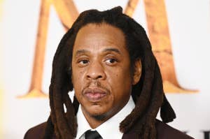 Jay-Z with shoulder-length dreadlocks wears a suit and tie, attending an event with a patterned backdrop
