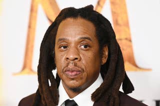 Jay-Z with shoulder-length dreadlocks wears a suit and tie, attending an event with a patterned backdrop