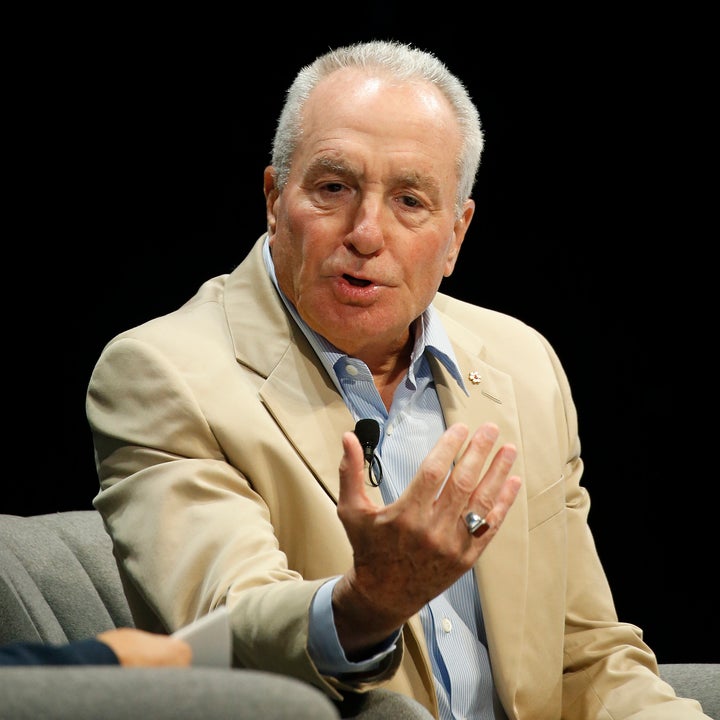 Lorne Michaels in a suit speaks animatedly while seated on a stage, gesturing with their hand