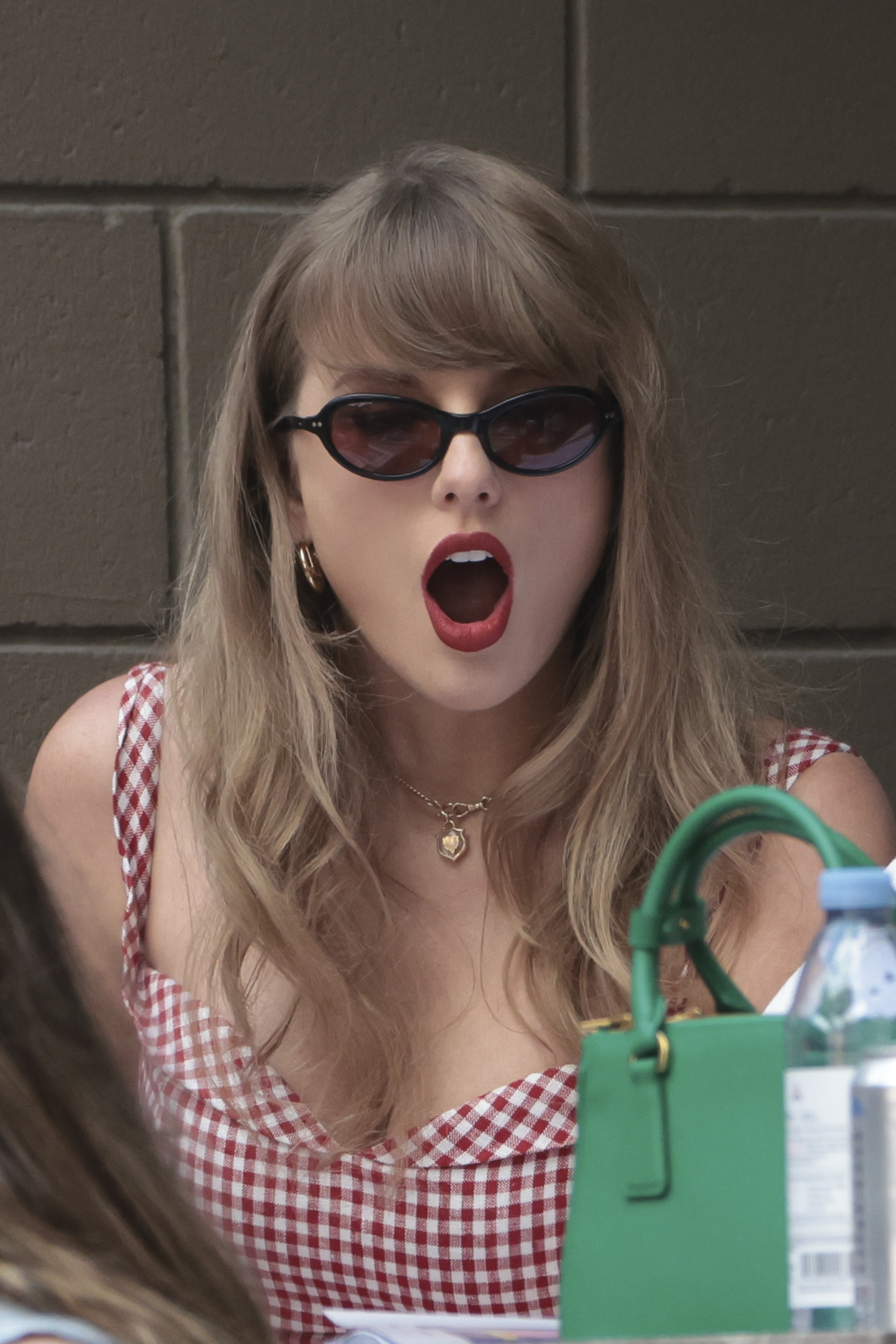 Taylor Swift in a gingham dress with sunglasses, surprised expression, sitting outdoors with a handbag visible