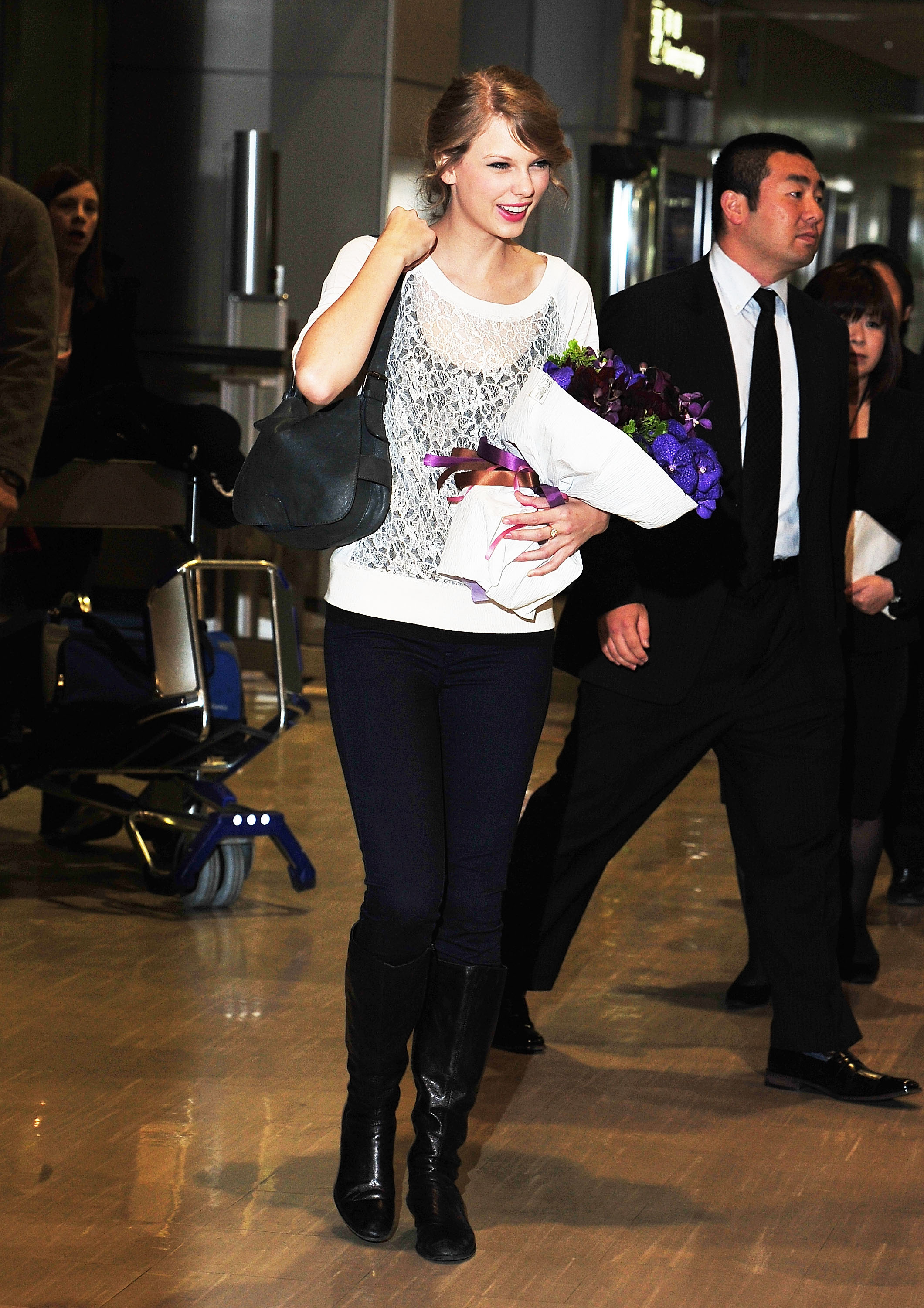 Taylor Swift at an event holding a bouquet, wearing a lace knit top, dark jeans, and knee-high boots, smiling while walking with a shoulder bag