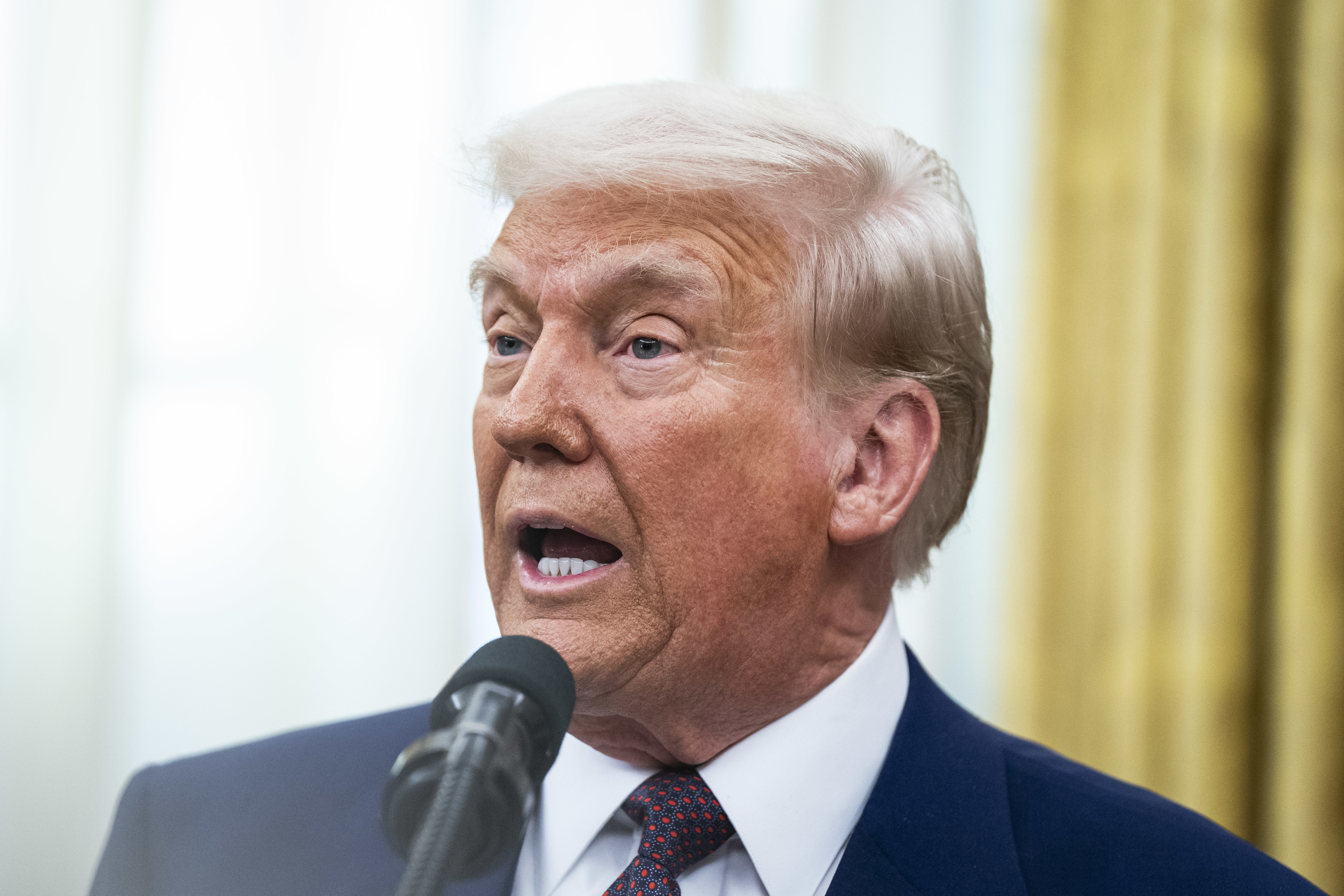 Donald Trump speaking at a podium with a microphone, wearing a dark suit and white shirt, in an indoor setting.