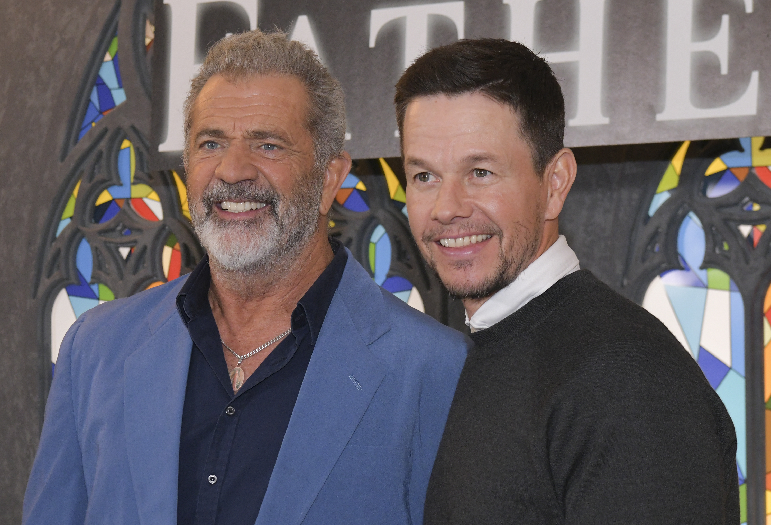 Mark Wahlberg & Mel Gibson smiling at an event. One wears a blue suit, the other a dark sweater over a white shirt. Stained glass-style decor in the background