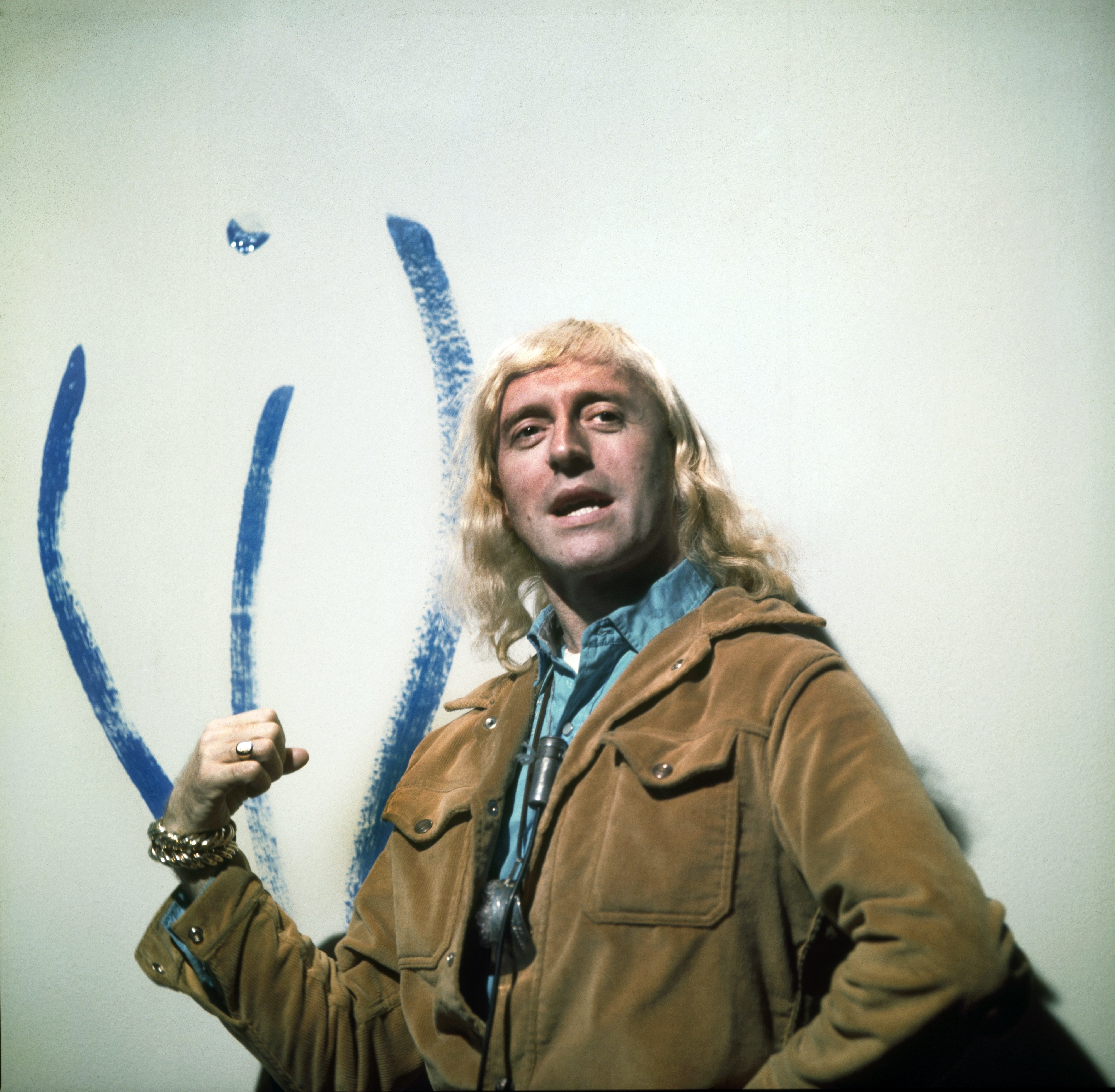 Jimmy Savile with long hair wearing a suede jacket and holding a paintbrush, stands in front of a large abstract blue painting
