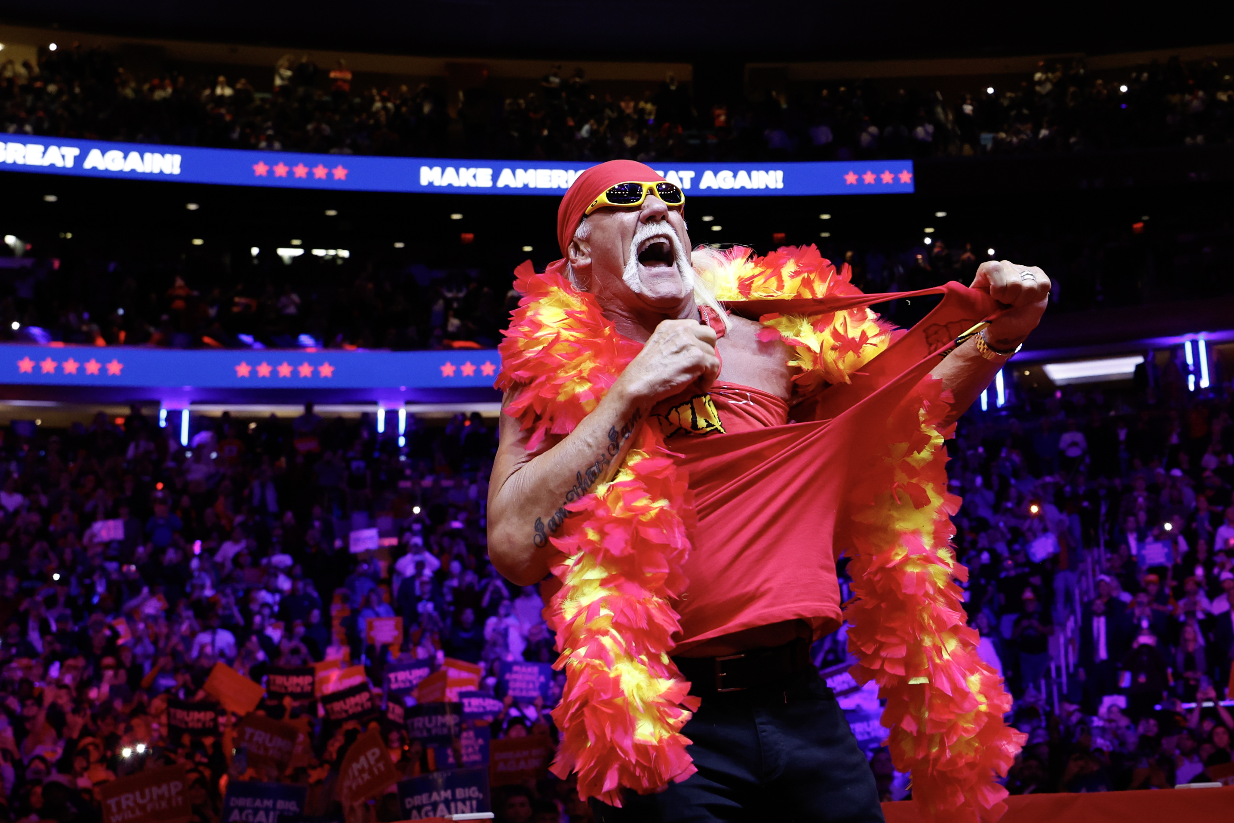 Hulk Hogan on stage wearing a feather boa and tearing a red shirt, surrounded by a cheering crowd at an event.