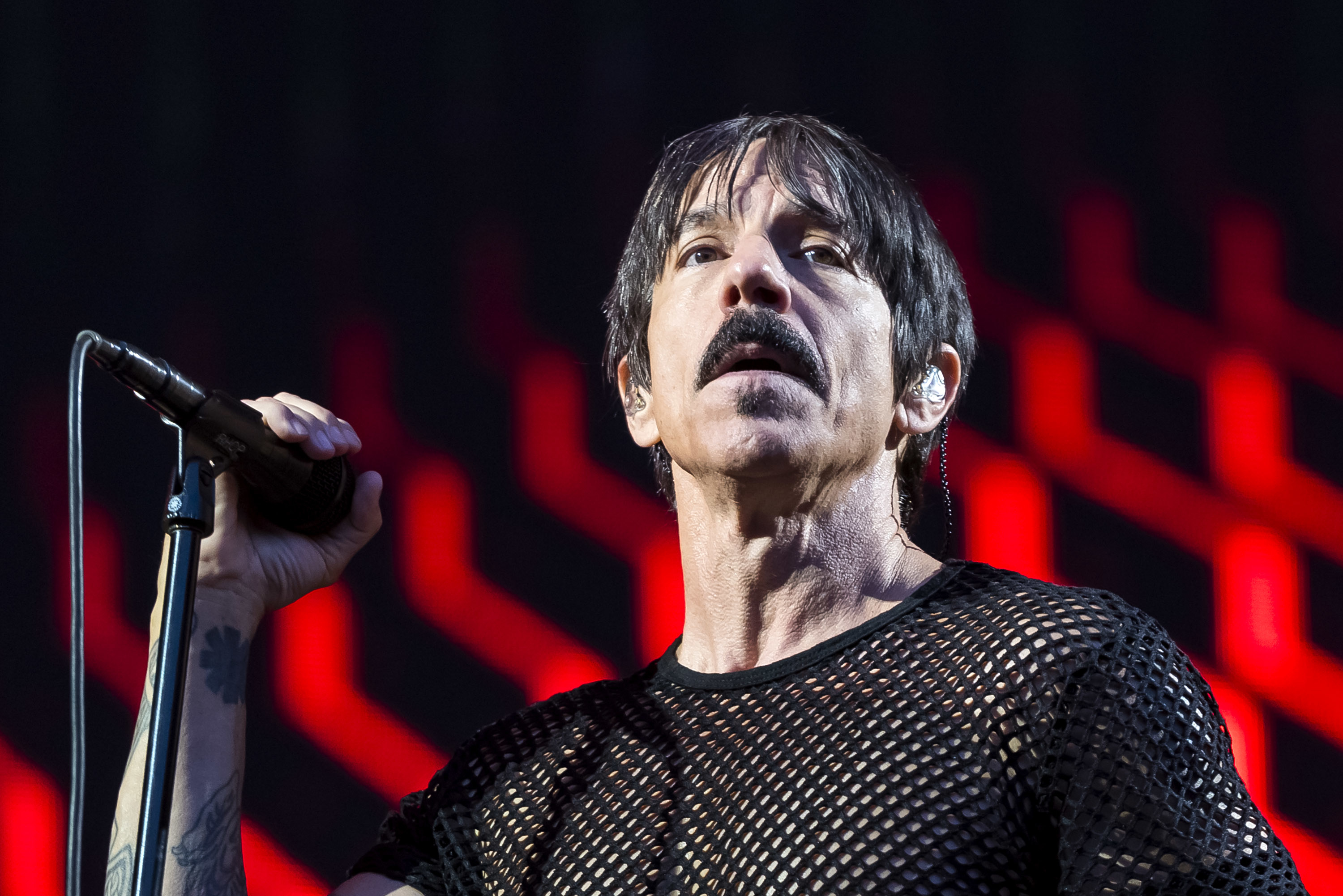 Anthony Kiedis singing on stage, wearing a black mesh shirt, holding a microphone, with red geometric background lights