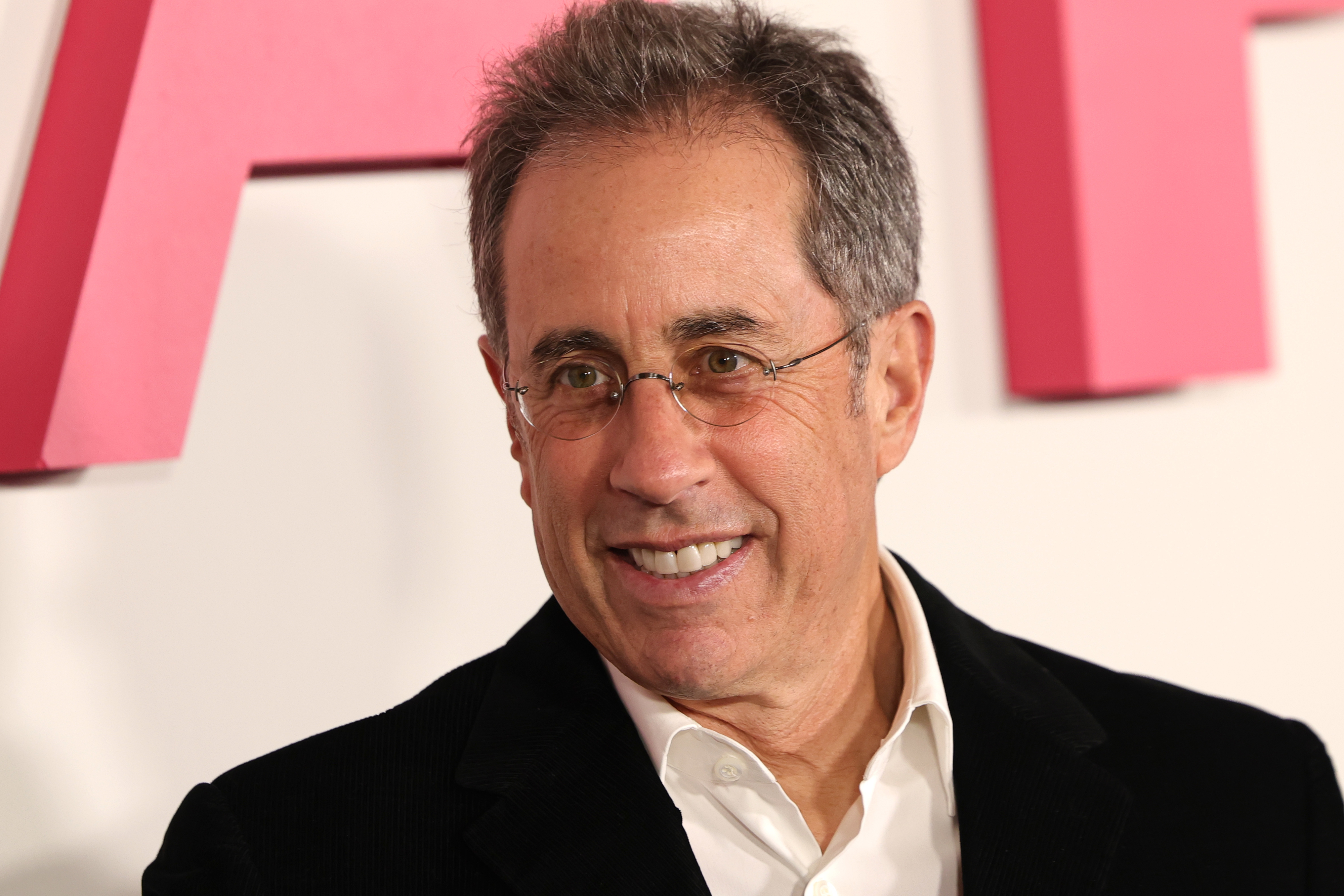 Jerry Seinfeld in a suit with glasses smiles at an indoor event.