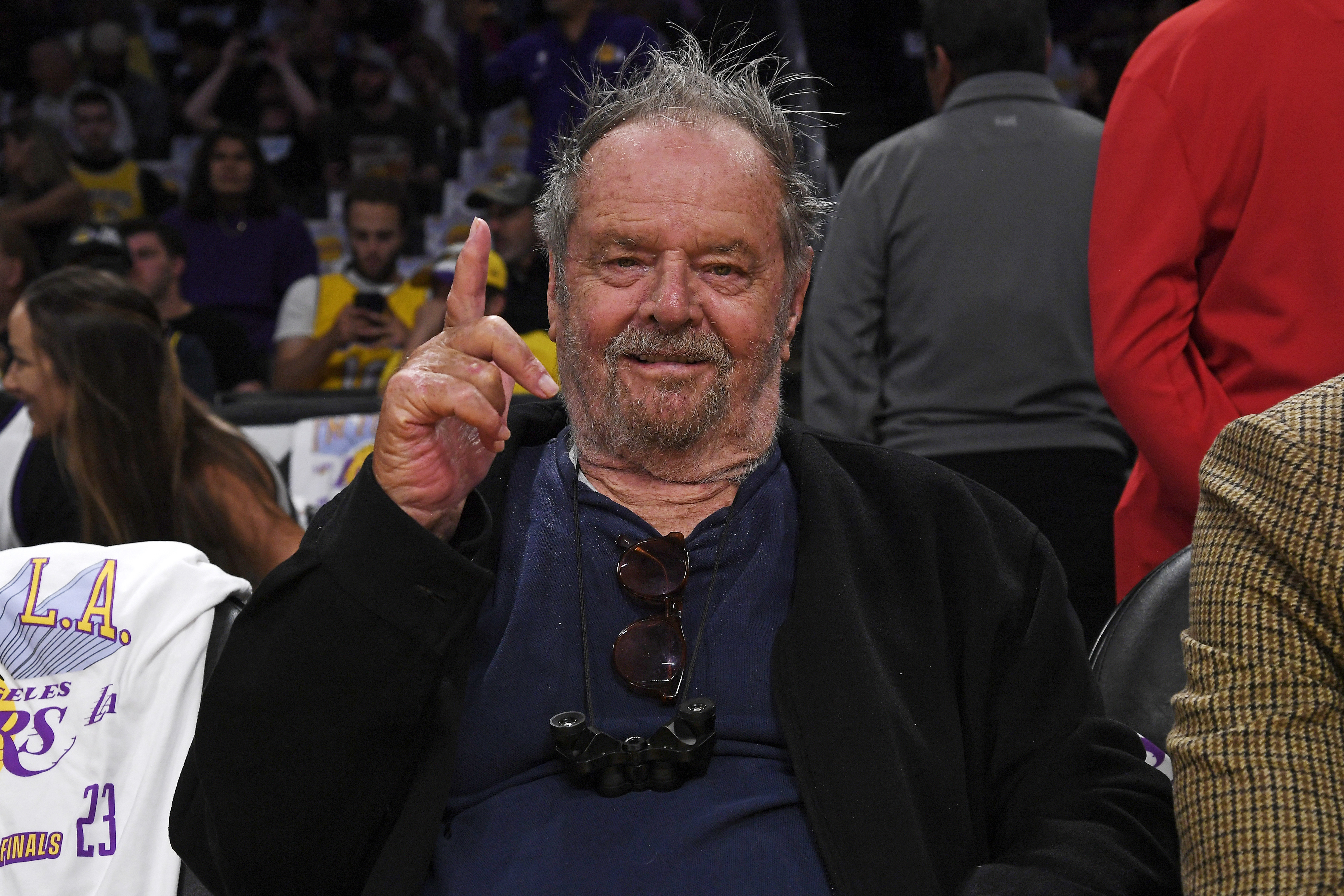 Jack Nicholson courtside at a basketball game, wearing a jacket and holding up one finger.