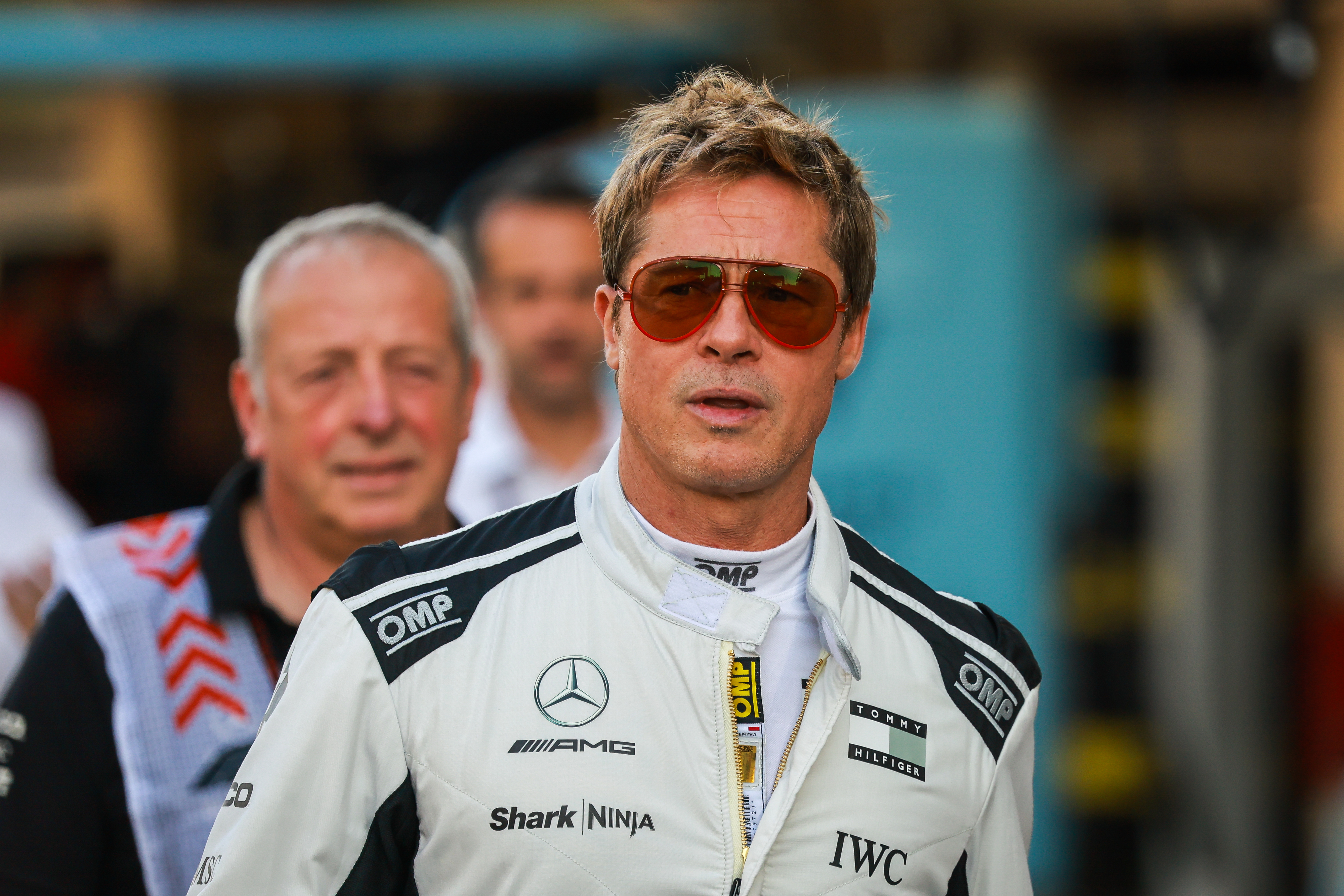 Brad PItt in a racing suit with sponsors' logos, wearing sunglasses, stands in a professional setting. Another person is blurred in the background