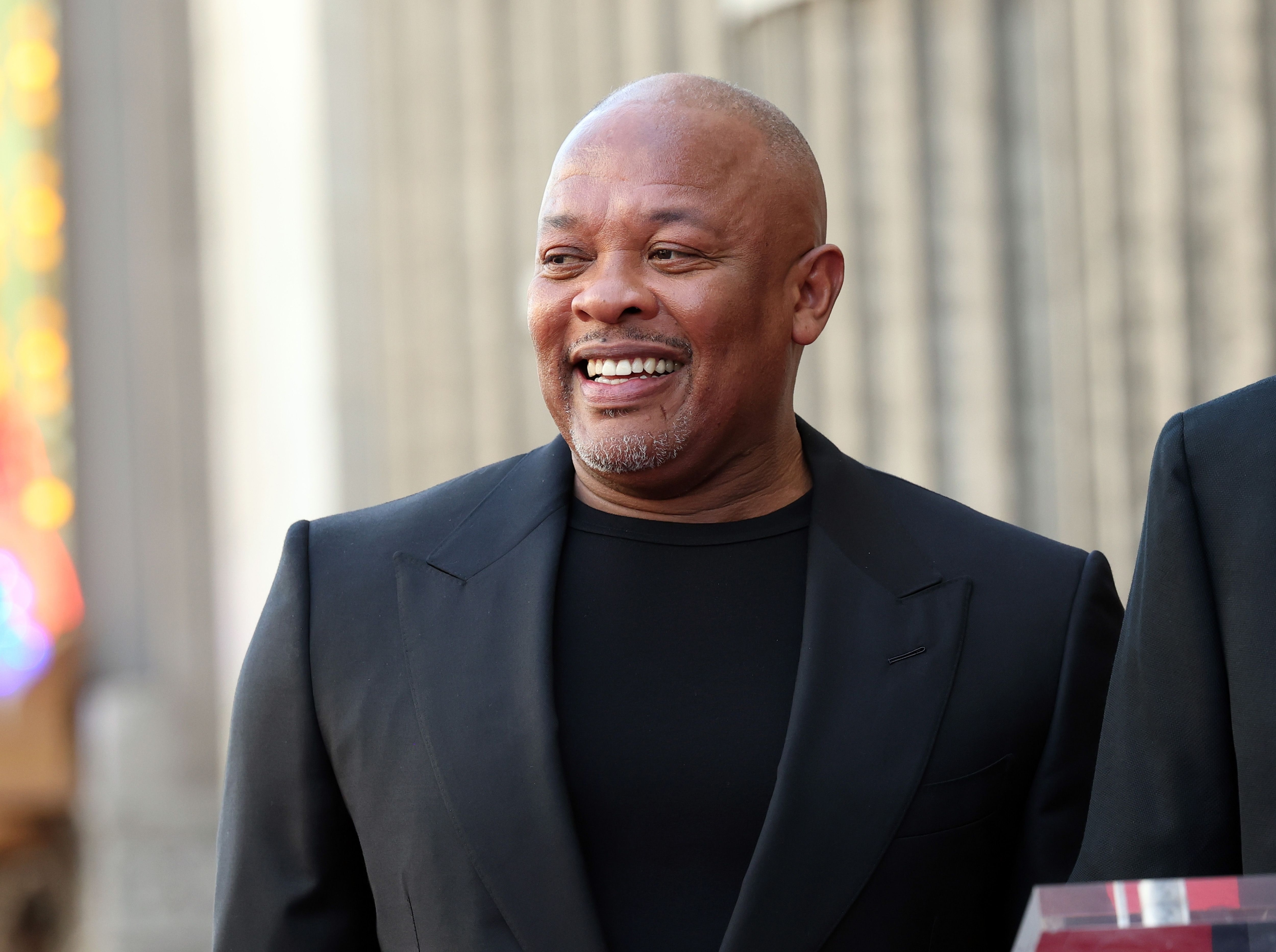 Dr. Dre in a black suit jacket stands outdoors in front of a building