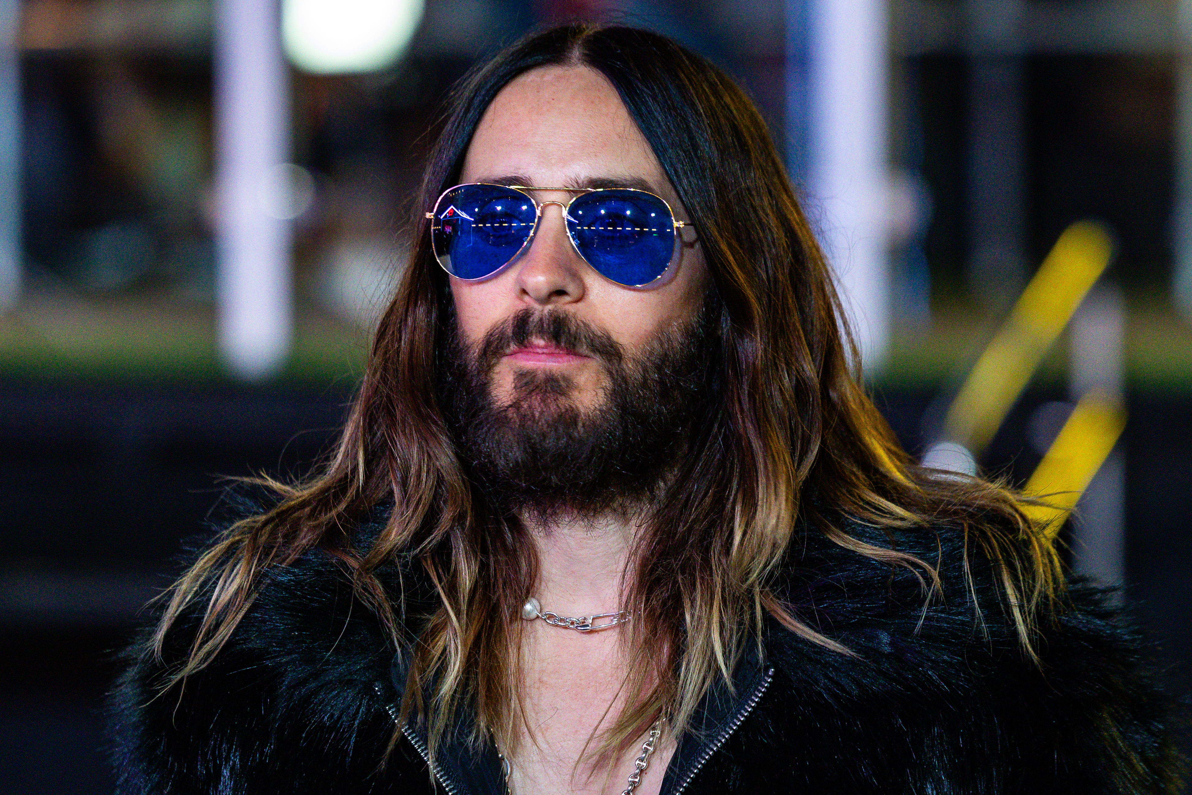 Jared Leto with long hair and beard wearing sunglasses and a dark, furry jacket, standing outdoors
