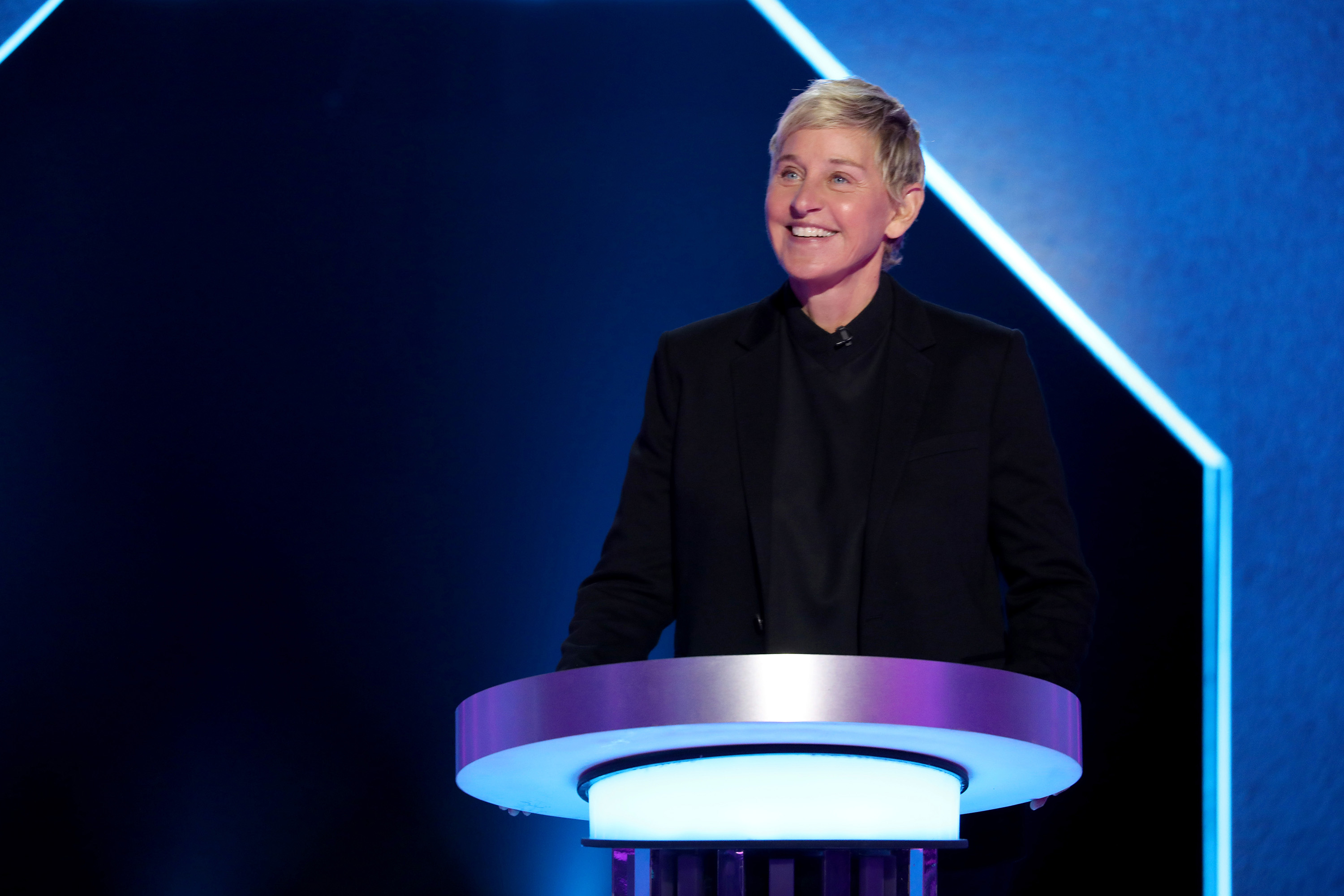 Ellen Degeneres in a dark suit stands smiling behind a podium on a brightly lit stage