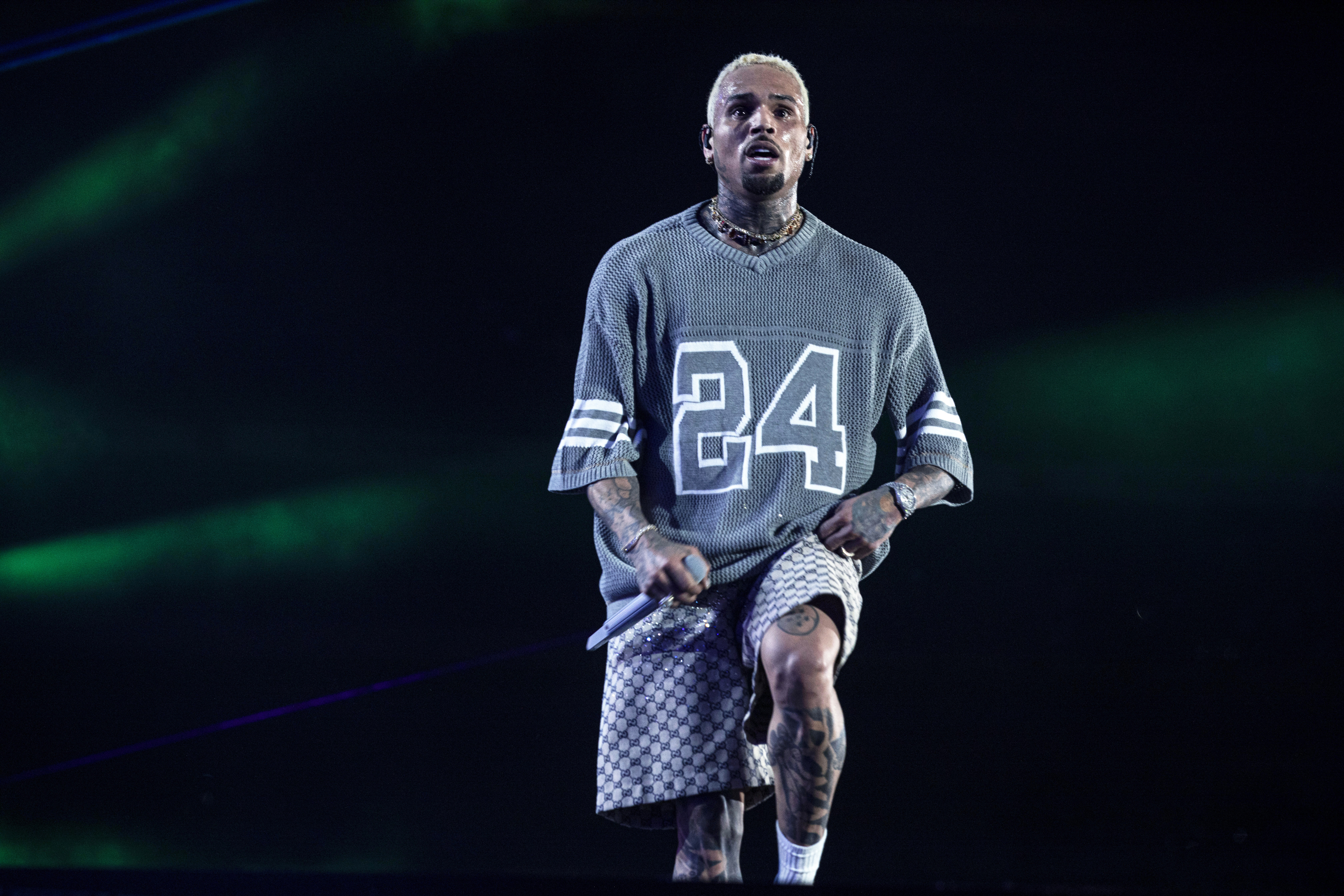 Chris Brown performing on stage, wearing a sports-style jersey with the number 24 and checkered shorts. They are holding a microphone