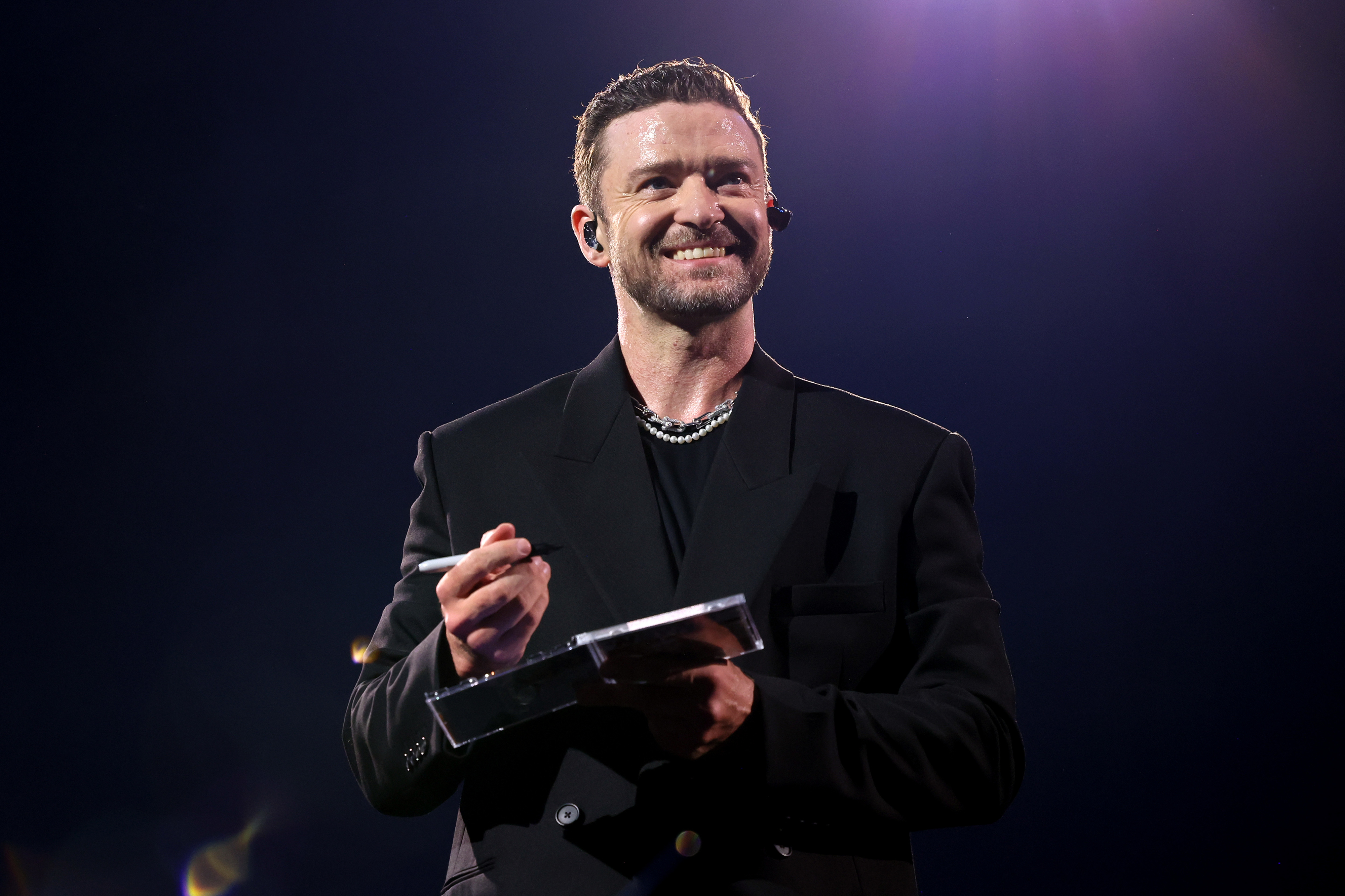A person on stage holds a microphone and a notepad, wearing a dark blazer and shirt, smiling at the audience