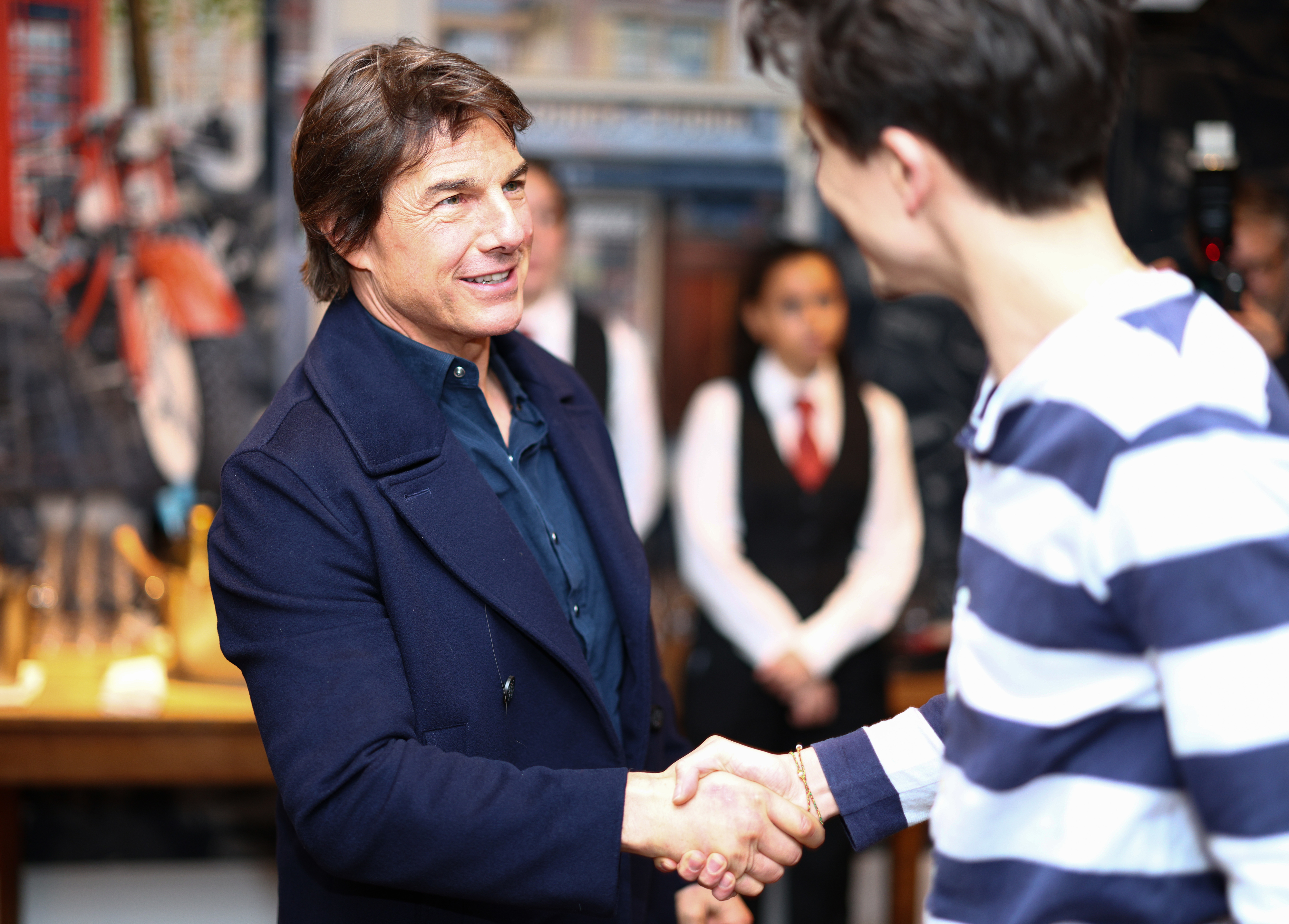 Tom Cruise In a dark coat shakes hands with another person in a striped shirt at a gathering.