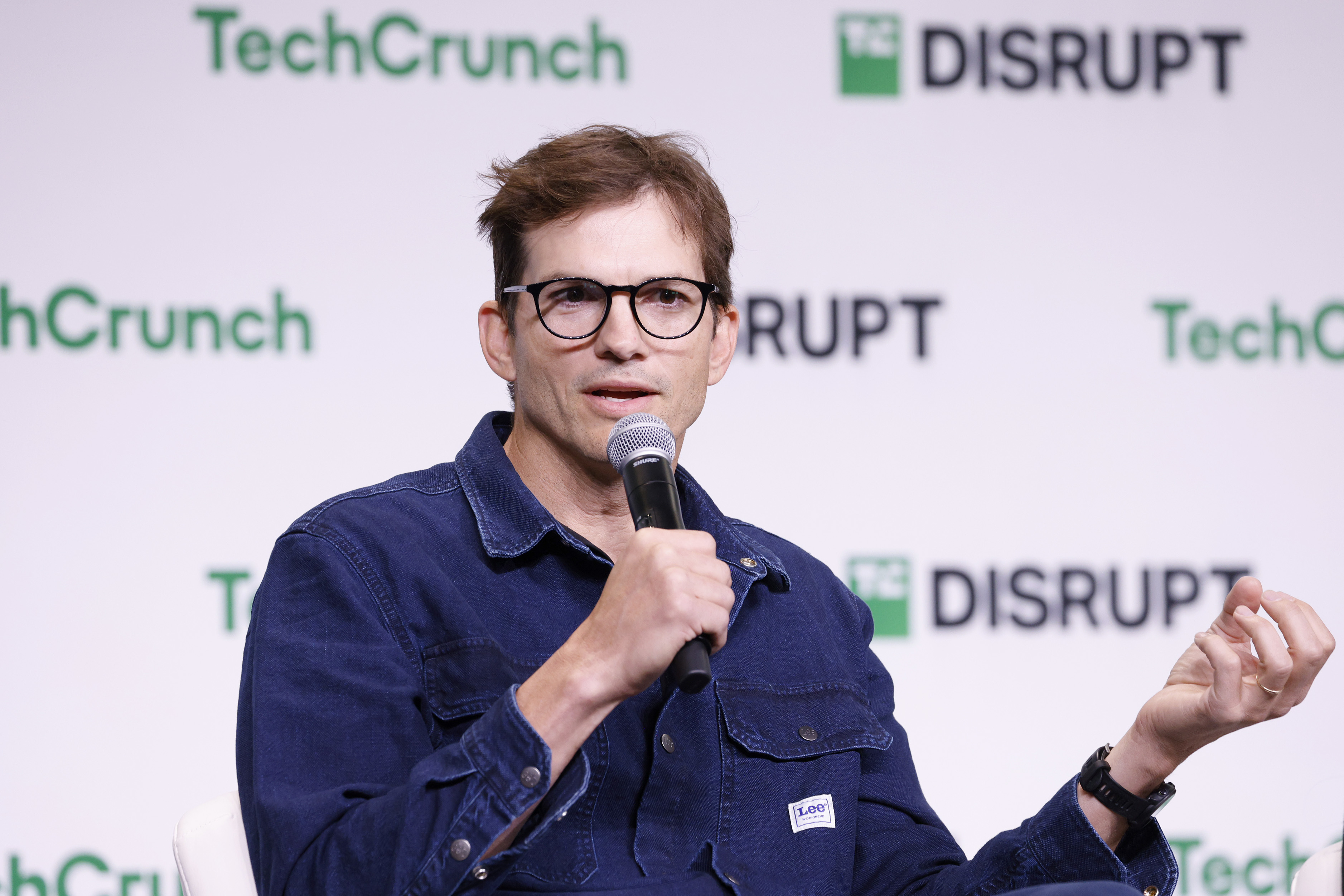 Ashton Kutcher wearing glasses and a denim shirt speaks into a microphone at a TechCrunch Disrupt event