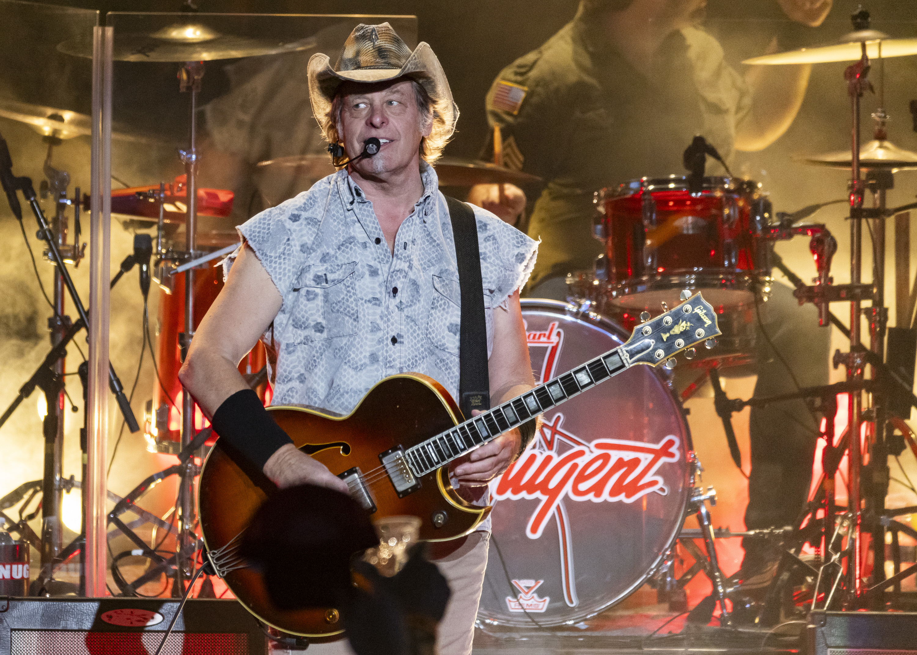 Ted Nugent on stage with guitar, wearing a sleeveless patterned shirt and cowboy hat, performing in front of a drum set