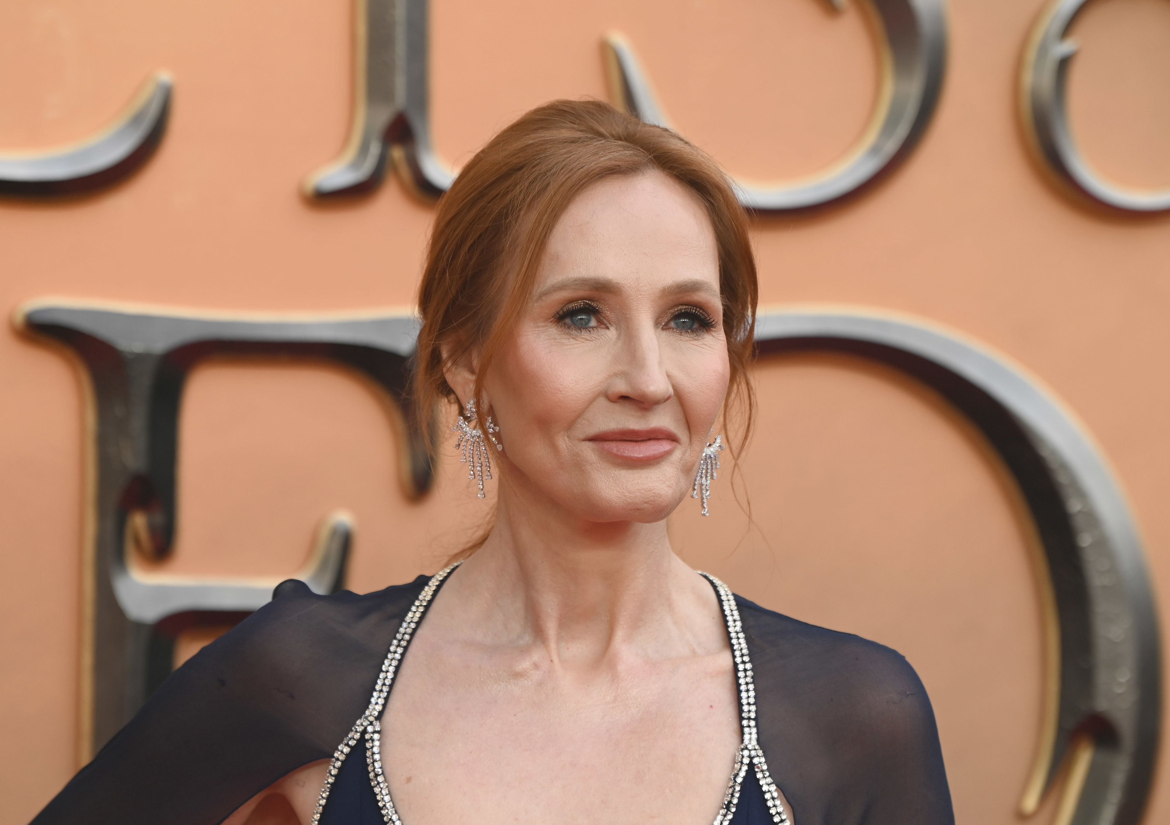 JK Rowling on a red carpet wearing a sheer-sleeved dress with sparkling trim and drop earrings, standing in front of a large, metallic text backdrop