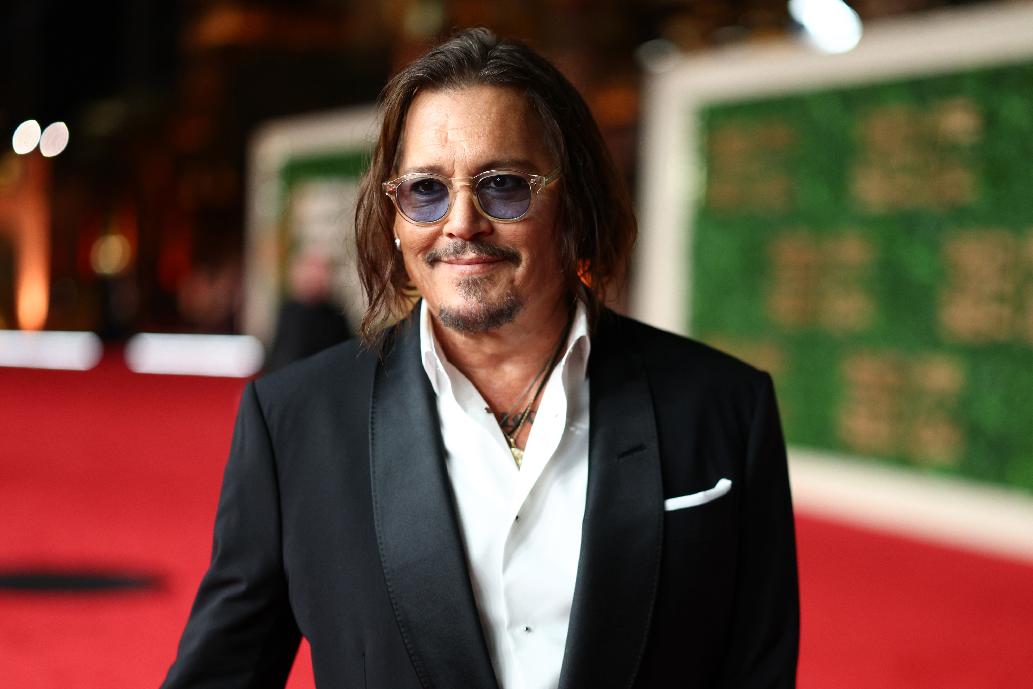 Johnny Depp in a tailored black suit and open-collared white shirt on a red carpet. They have medium-length hair and sunglasses