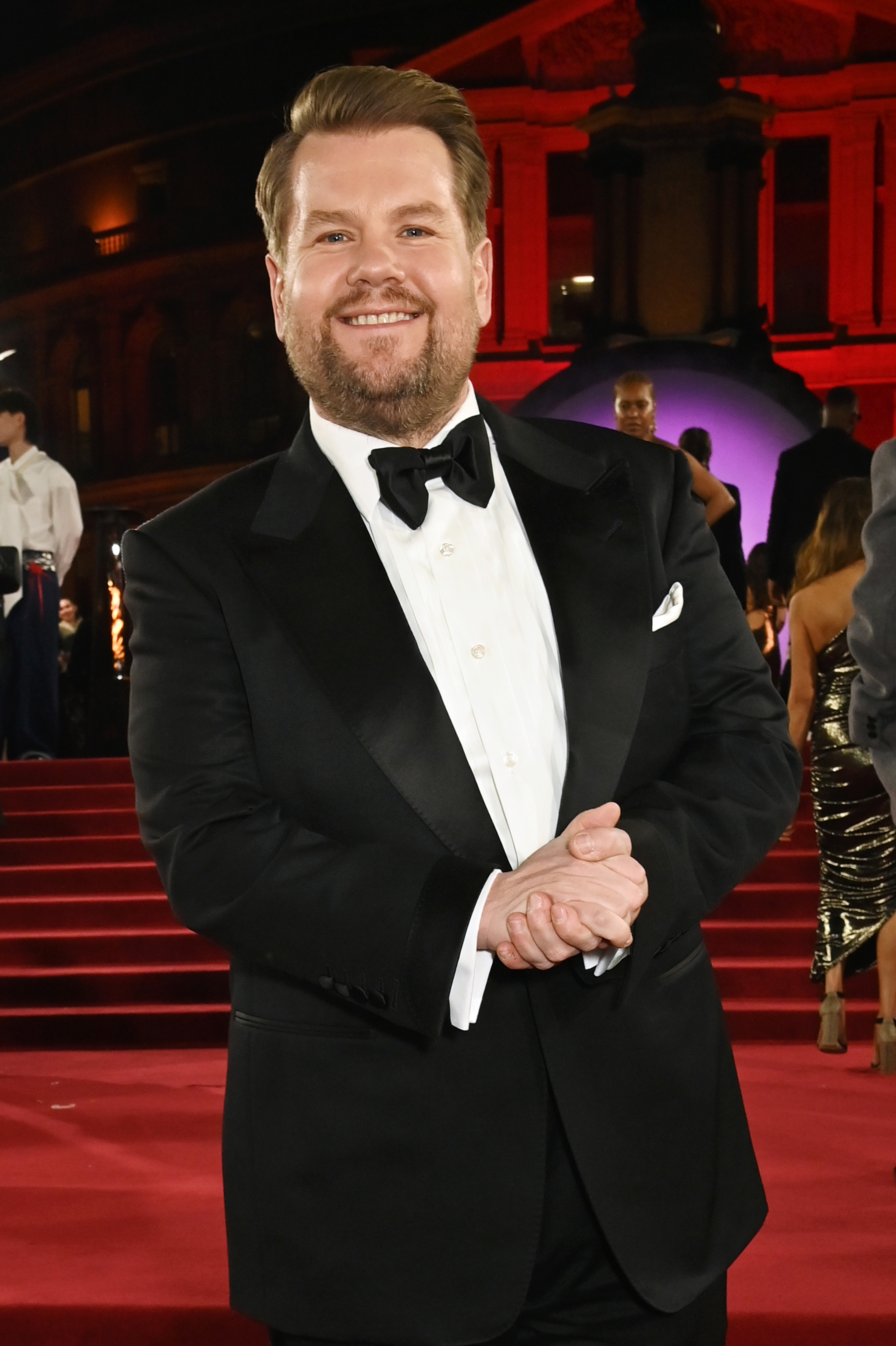 James Corden in a black tuxedo with a bow tie on a red carpet, smiling with clasped hands