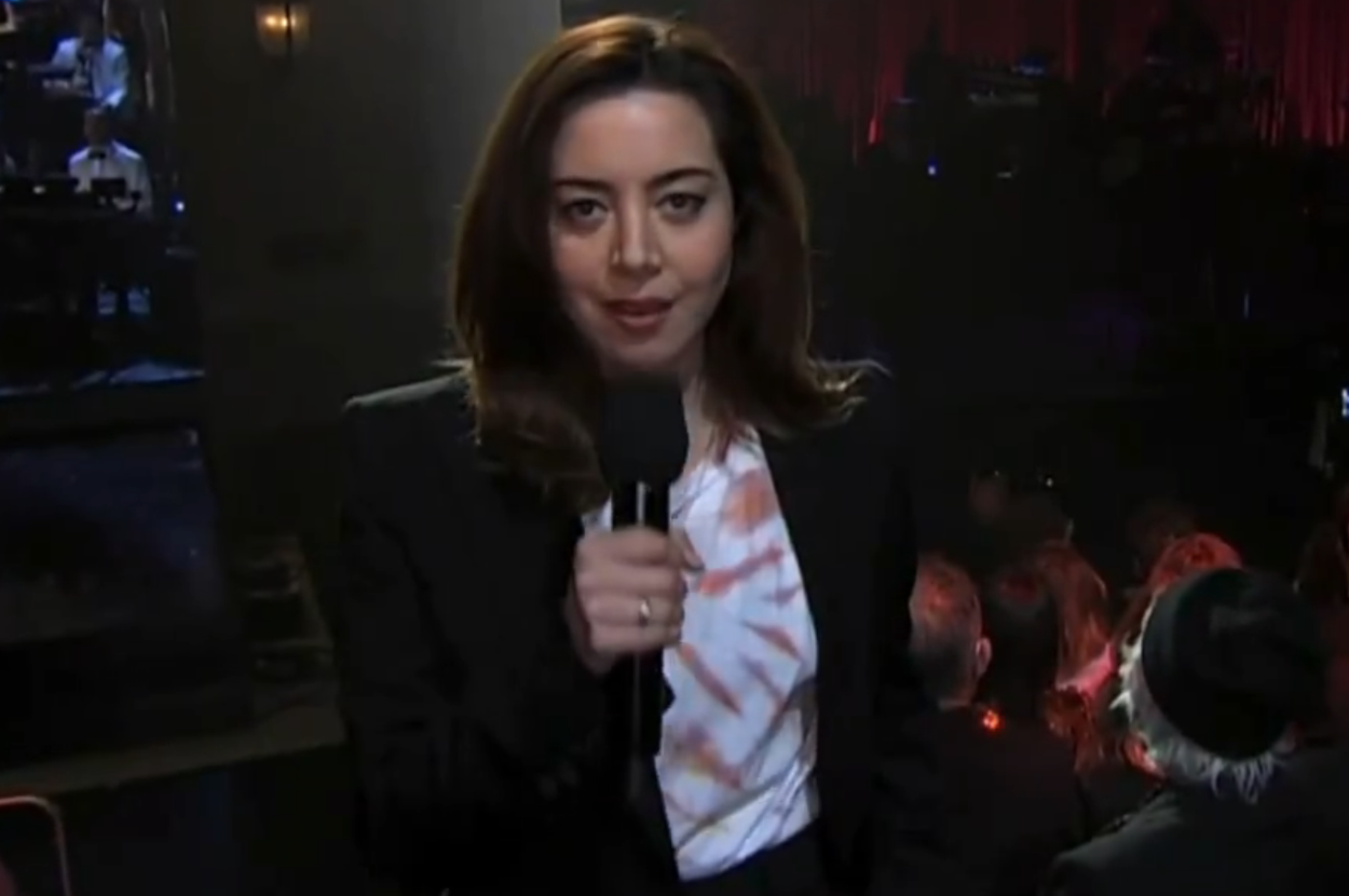 Aubrey Plaza holding a microphone on stage, wearing a suit with a tie-dye shirt, addressing an audience
