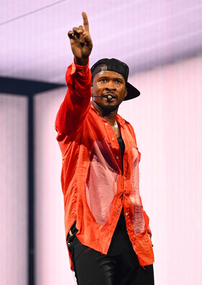 Usher on stage wearing a loose shirt and cap, holding a microphone and gesturing upward with one finger raised