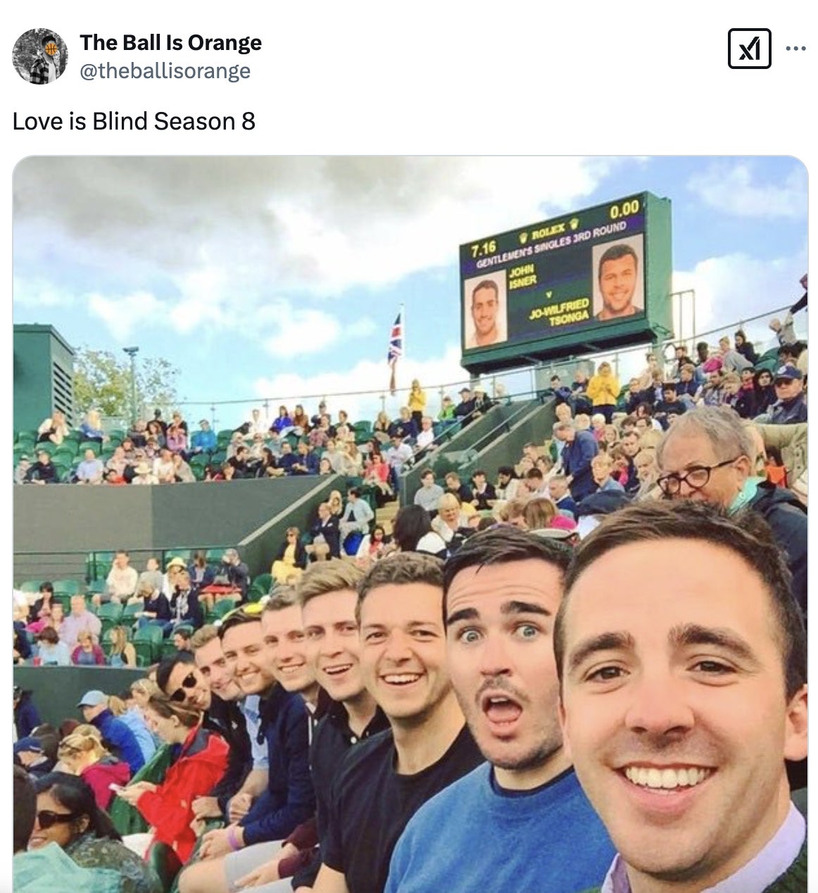 Group of eight men taking a selfie at a tennis match, with a scoreboard visible in the background